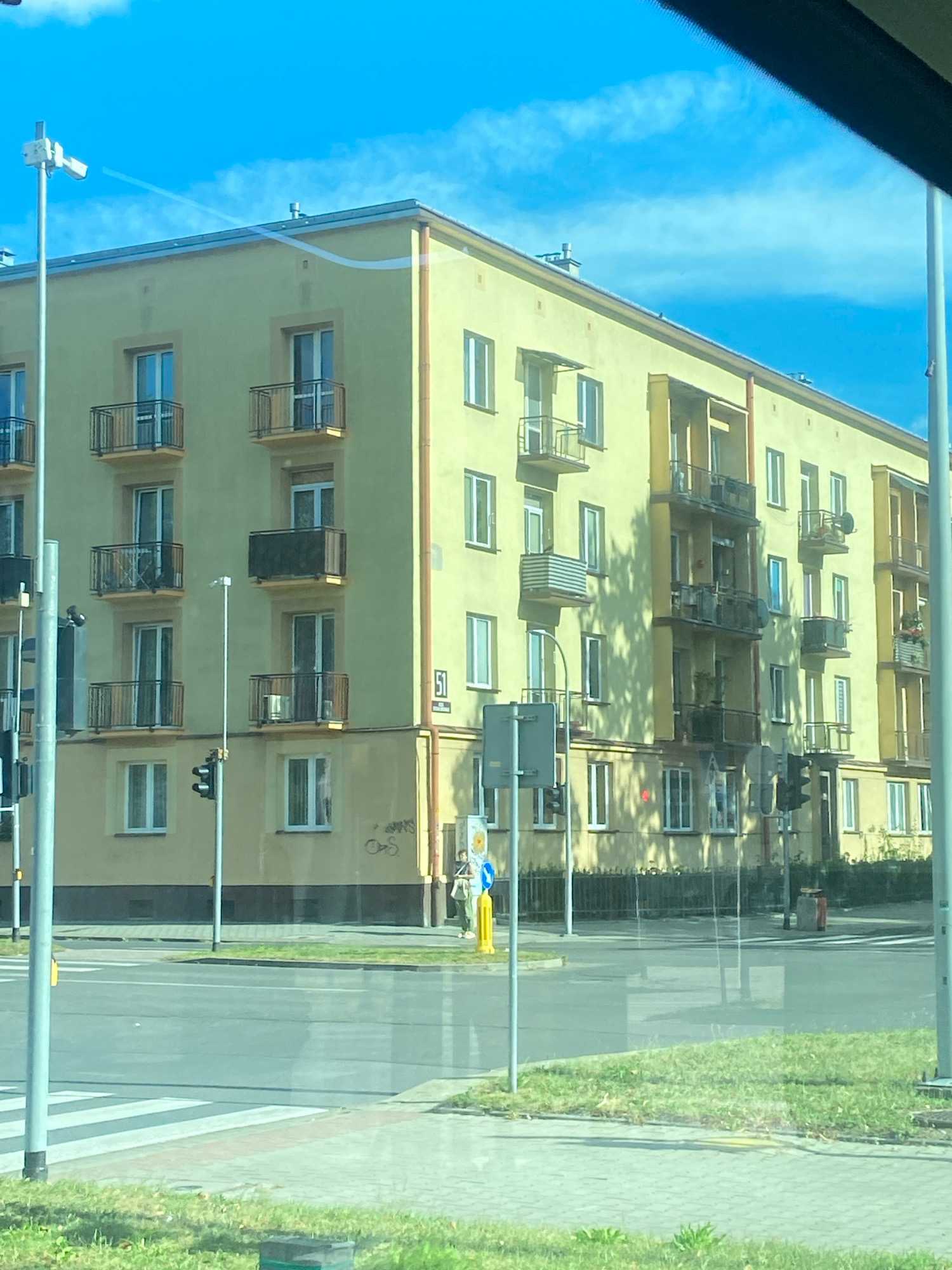 This is an eye-level, medium-shot photograph of a yellow apartment building located in Nowy Sącz, Poland. The building is four stories tall with multiple balconies on each floor. The sky above is a clear, bright blue with a few wispy clouds. In the foreground, a pedestrian crossing is visible on the road, indicating an intersection. Traffic lights and streetlights are present, and a person is walking on the sidewalk. The building appears to be a residential complex, and the surrounding environment suggests a typical urban setting. The overall impression is of a sunny day in a European city.