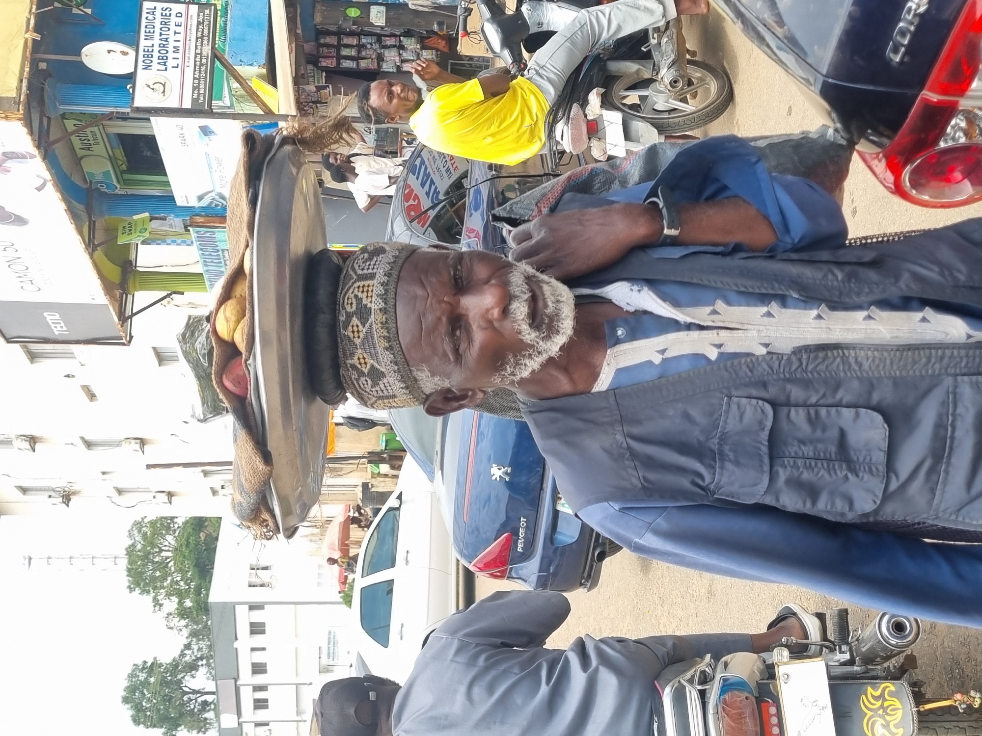 A man with a gray beard and wearing a blue vest over a blue and white patterned garment carries a large tray of goods on his head. He is standing in a busy street scene in Jos, Nigeria. The man is looking directly at the camera. In the background, a white car and a blue Peugeot are visible, along with motorcycles and various shops lining the street. One shop has a sign that reads "NOBEL MEDICAL LABORATORIES LIMITED." The overall atmosphere suggests a vibrant market or commercial area in the city.