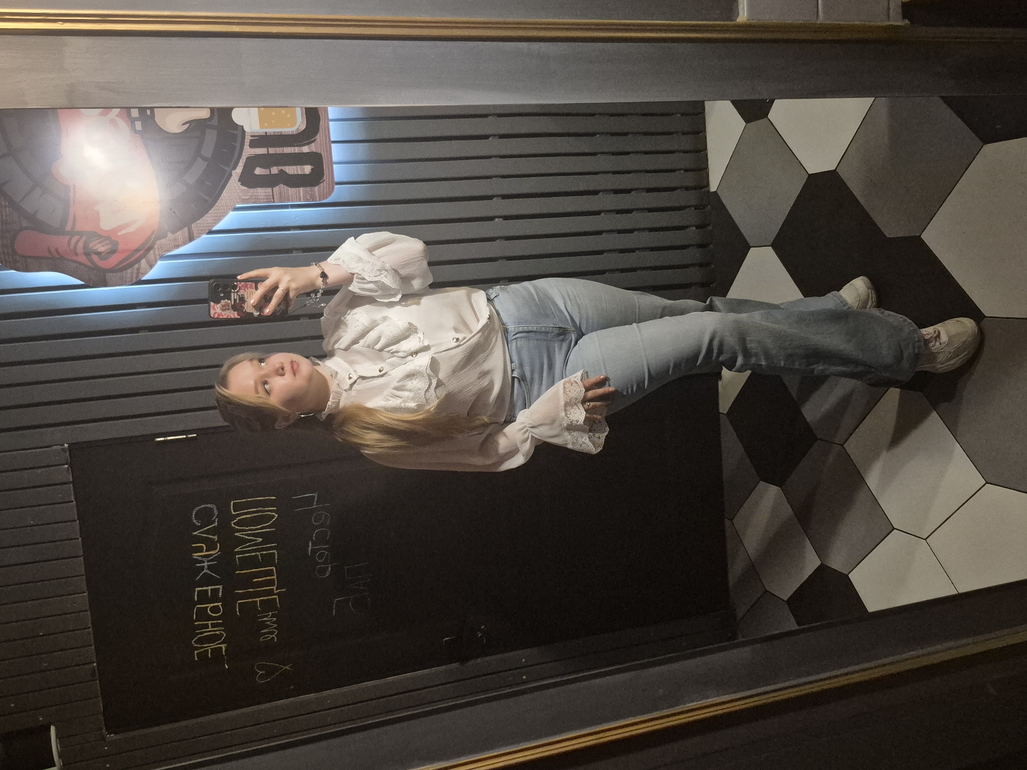 A young woman stands in a restroom, taking a selfie in a mirror. She has blonde hair pulled back in a ponytail and is wearing a white ruffled blouse with lace detailing and light-wash flared jeans. Her footwear appears to be light-colored sneakers. The restroom has a dark, vertical panel wall and a black door with writing on it, which appears to be in Cyrillic script. The floor is tiled in a geometric pattern of black, white, and grey hexagons. To the right of the woman, a neon sign depicting a cartoonish figure holding a mug is visible in the mirror reflection. The writing on the door, when deciphered, suggests the location is in Chaikovsky, Russia, possibly a bar or establishment with a playful atmosphere. The overall ambiance of the scene is casual and slightly dimly lit.