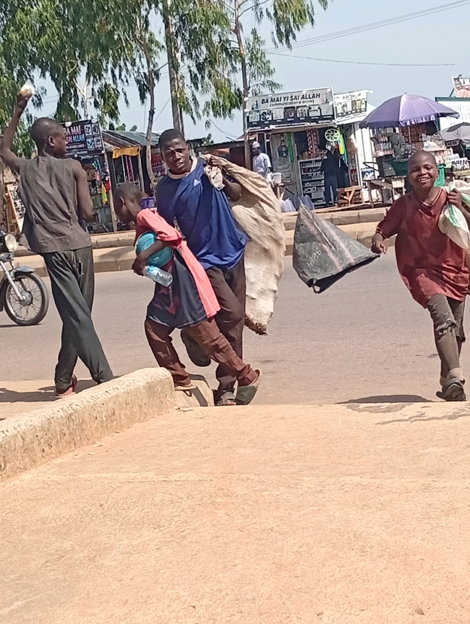 In Tumfure, Nigeria, a group of individuals, appearing to be children and young men, are walking on a dusty street. The foremost person on the right, a boy in a reddish-brown shirt, smiles broadly as he carries a white plastic bag. To his left, a young man in a blue shirt is carrying a large, light-colored bundle over his shoulder. A younger child, wearing a pink and blue garment, clutches a blue ball and a water bottle. To the far left, a boy in a dark grey shirt is raising his arm. In the background, market stalls line the street, with various goods displayed. A purple umbrella provides shade over one of the stalls, and a motorcycle is parked to the left. The scene is illuminated by bright sunlight, casting shadows on the ground.