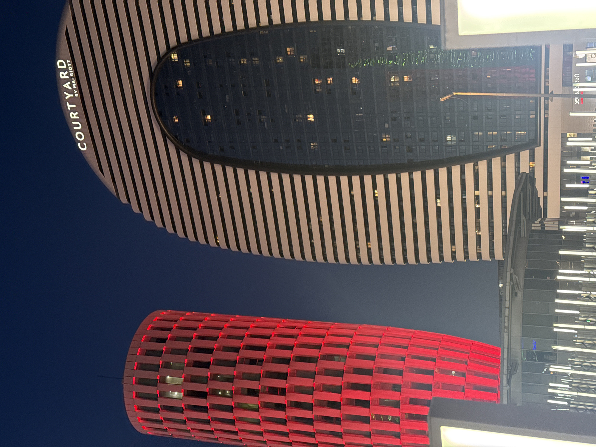 At night, the Courtyard by Marriott hotel in Batumi, Georgia, stands with its distinctive black and white striped facade. Beneath it, a cylindrical building with red rectangular segments emits a bright glow. Further in the background, another building with a glass facade reflects the city lights. The sky above is a dark blue.