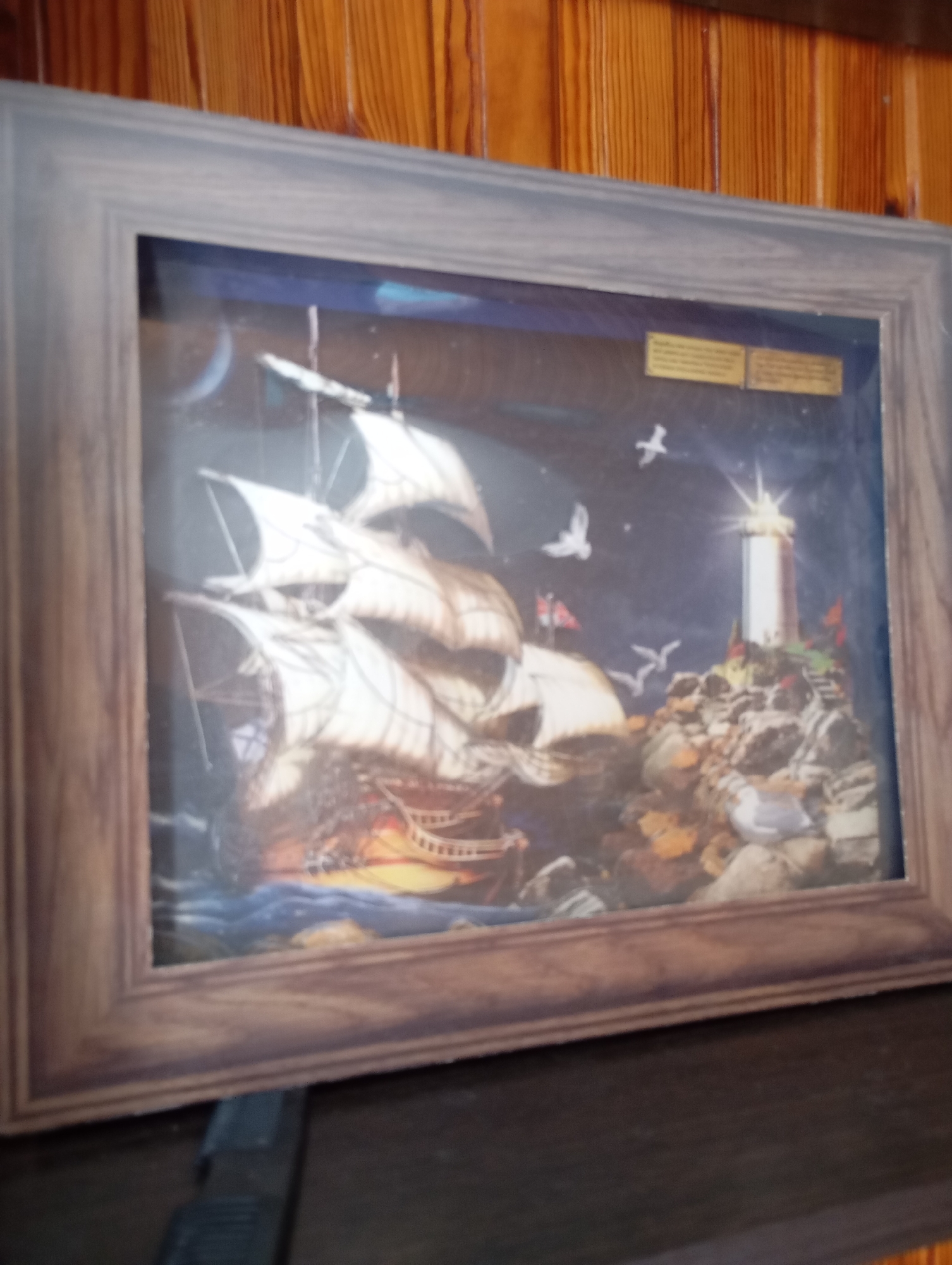 A framed shadow box diorama depicts a scene with a tall ship sailing on dark, wavy water under a night sky. A lighthouse stands on a rocky outcrop to the right, emitting a bright light. Several white birds are in flight around the ship and lighthouse. The diorama is framed in a wooden border and rests on a dark surface. The background appears to be a wooden wall. The location in the city of Льниха, Russia is not discernible from the image.