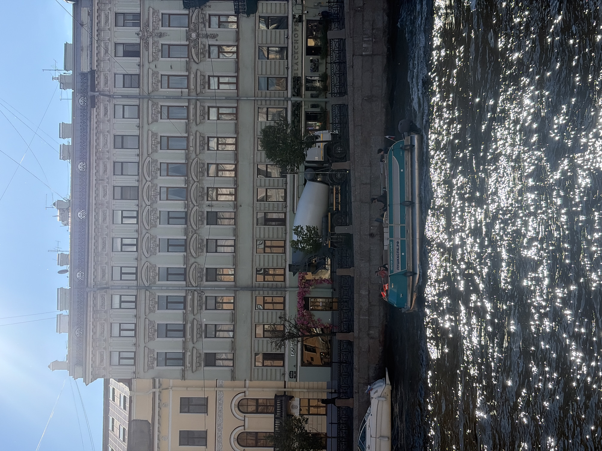 This media file captures a view of a canal in Saint Petersburg, Russia, on a sunny day. The scene is dominated by the architecture of a building lining the canal, featuring multiple stories with numerous windows and decorative elements. Sunlight glints off the water's surface, creating a shimmering effect.
Several objects are visible along the canal bank and in the water. A concrete mixer truck is parked on the embankment, suggesting potential construction or maintenance work. A green and white boat, possibly a tour boat, is docked in the canal, with at least two individuals visible on board, one wearing an orange life vest. Another white boat is also present, partially visible at the bottom of the frame.
The building's facade exhibits a light green color and intricate architectural details, including moldings and sculptures around the windows. The time of day appears to be afternoon, given the bright sunlight and the angle of the shadows. The weather is clear and sunny.
On the building, text is visible on a sign that reads "BLACKCHOPS". Additionally, text on the green and white boat appears to be in Cyrillic script.
The overall impression is one of a picturesque urban scene in Saint Petersburg, blending architectural heritage with the activity of the canal.