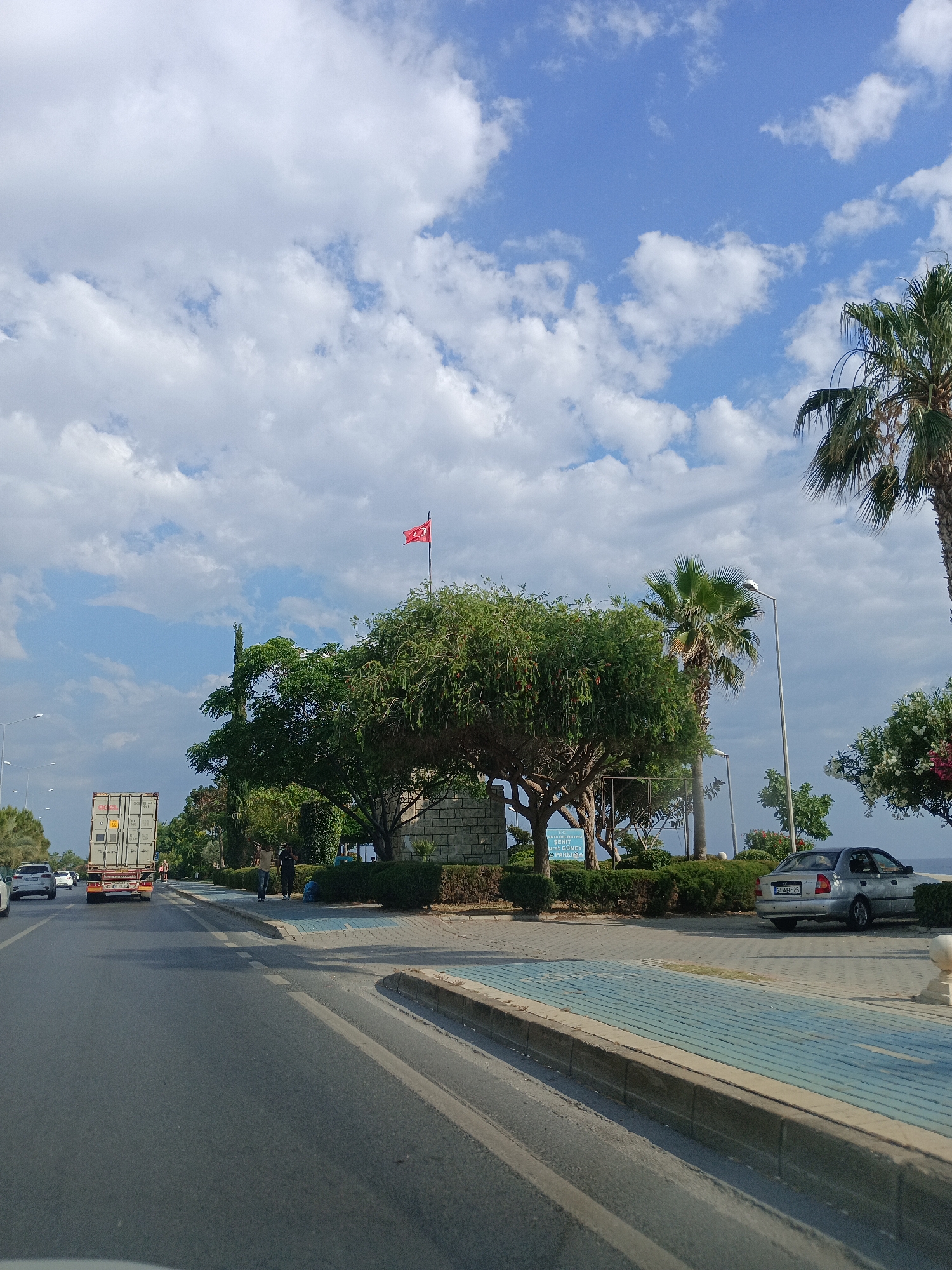 The image depicts a sunny day in Alanya, Turkey, with a bright blue sky dotted with fluffy white clouds. The scene is a coastal road lined with palm trees and lush green foliage. In the foreground, a paved road leads into the distance, with a curb and a blue-tiled walkway on the right.
A large white shipping container is being transported on the back of a truck, moving along the road. Two men are walking on the sidewalk, appearing to be in casual conversation. Further down the road, a silver car is parked. A Turkish flag flutters proudly on a flagpole atop a stone structure.
The presence of palm trees, the Turkish flag, and the Mediterranean Sea visible in the background strongly suggest the location is Alanya, Turkey. The time of day appears to be late morning or early afternoon, given the bright sunlight and clear sky. The overall atmosphere is peaceful and leisurely, characteristic of a seaside resort town.
A sign is visible on the left side of the image, which reads in part: "ALANYA BELEDIYESI" and "GUNEY PARKING". This further confirms the location as Alanya, a municipality in Turkey, and indicates a parking area.