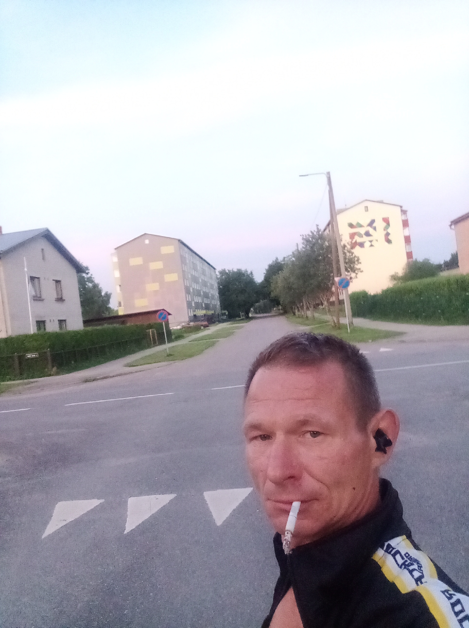 The image shows a man smoking a cigarette in Tapa, Estonia. He is wearing a black jacket with yellow and white accents. In the background, there are apartment buildings and trees. The sky is overcast. There are road markings in the foreground, indicating a street.