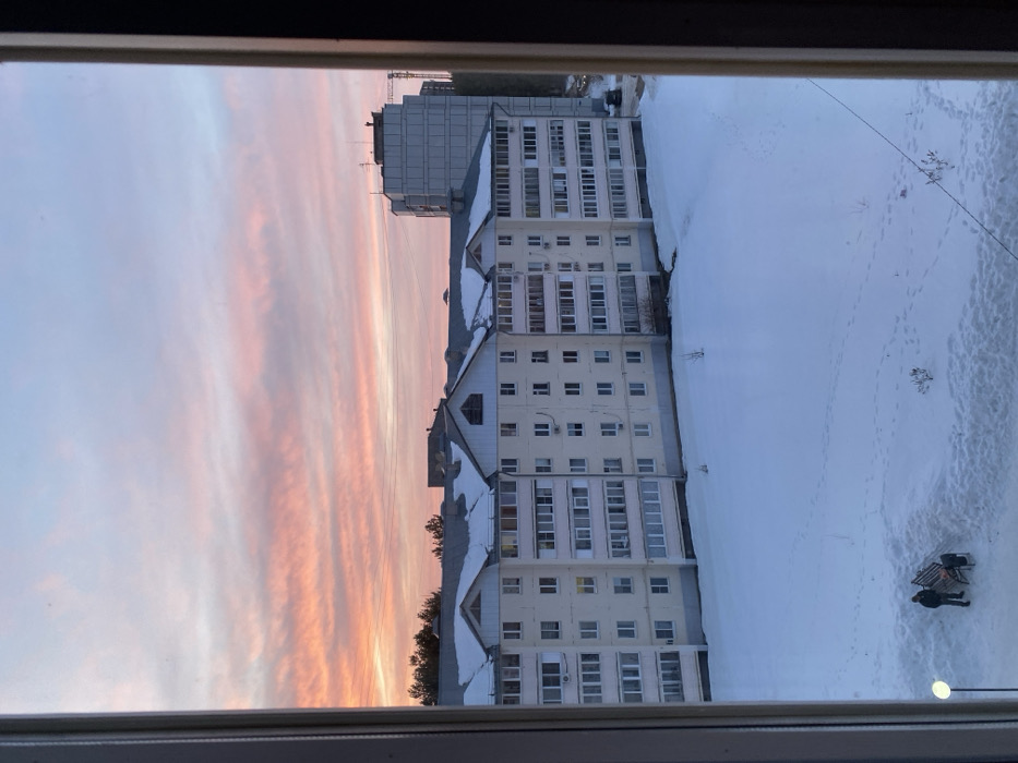 The image presents an urban winter scene in Chaikovsky, Russia, viewed through a window frame visible on the left and right. The upper half of the frame is dominated by a sky displaying vibrant pink and orange stratiform clouds against a lighter blue, indicative of dawn or dusk.
In the mid-ground, a multi-story, light-colored apartment building extends horizontally, featuring a complex roofline partially covered in snow. The building includes numerous rectangular windows, enclosed balconies, and several external air conditioning units. Behind it, to the right, a taller, grey, utilitarian building is visible, with a construction crane partially obscured further right. A line of evergreen trees appears on the distant horizon to the left.
The lower half of the image consists of a snow-covered ground plane, marked by multiple footprints. In the lower-left quadrant, two individuals are positioned near a dark, slatted park bench. One person stands to the left of the bench, while the other is seated on it; both are dressed in dark winter attire. A modern streetlight with a circular luminaire stands to the left of the individuals. Sparse, small bushes or saplings protrude from the snow. A dark vehicle is partially visible on the far right, on a cleared patch behind the apartment building.