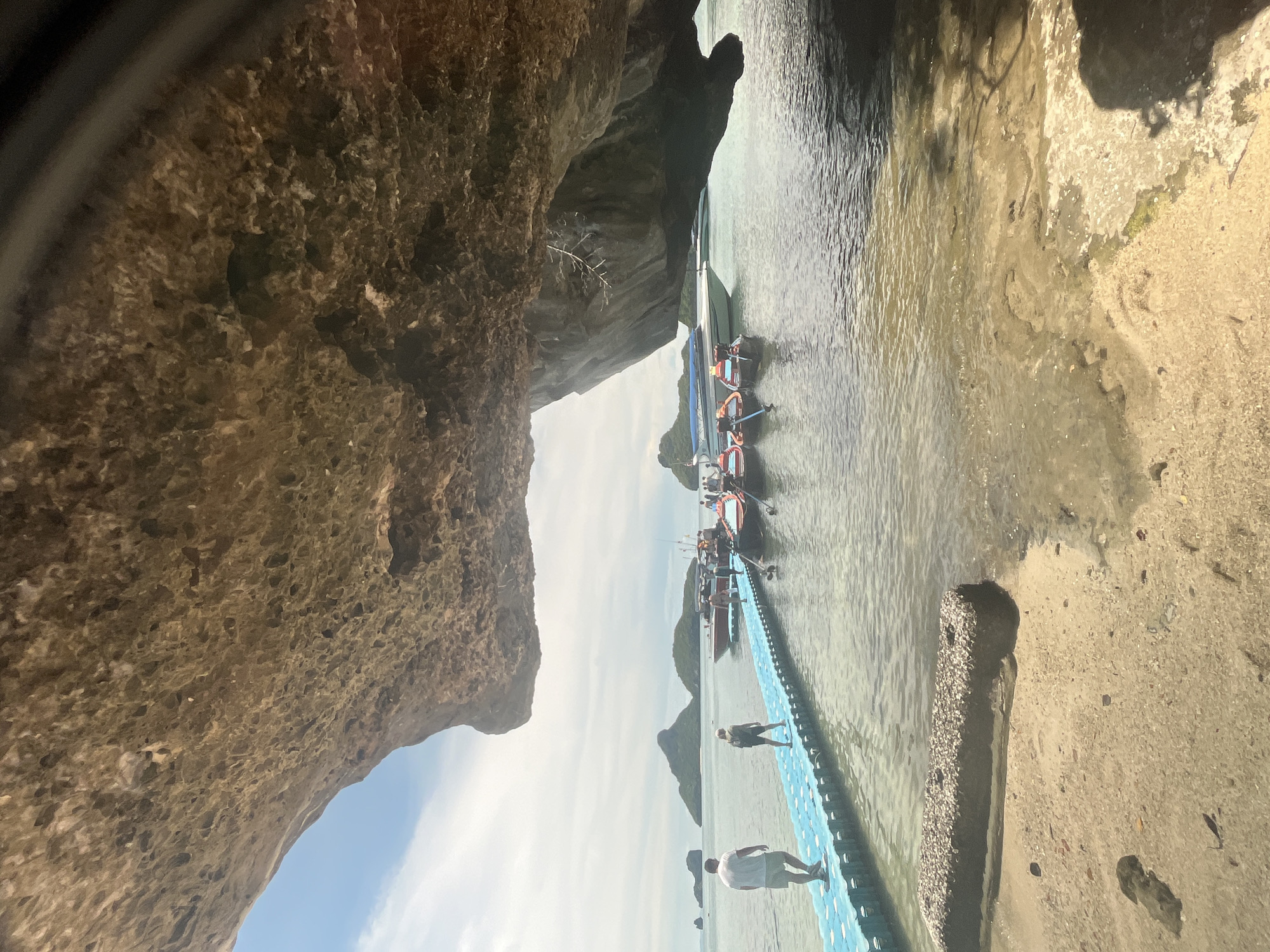 This is a photograph taken from underneath a large rock overhang, looking out at a tropical island scene. The overhang takes up the top portion of the image, creating a natural frame. In the middle ground, a blue floating walkway extends from a sandy beach into the clear, calm water. Two men are walking on the walkway away from the shore. Several small boats, some appearing to be longtail boats, are moored near the walkway, with people visible on them. In the background, a larger white boat is docked, and several lush, green islands rise from the sea under a cloudy sky. The water is shallow and clear near the shore, revealing sandy patches and some submerged rocks. The beach in the foreground is sandy with some scattered debris. The overall scene suggests a popular tourist location in Thailand, likely an island known for its karst topography and boat tours.