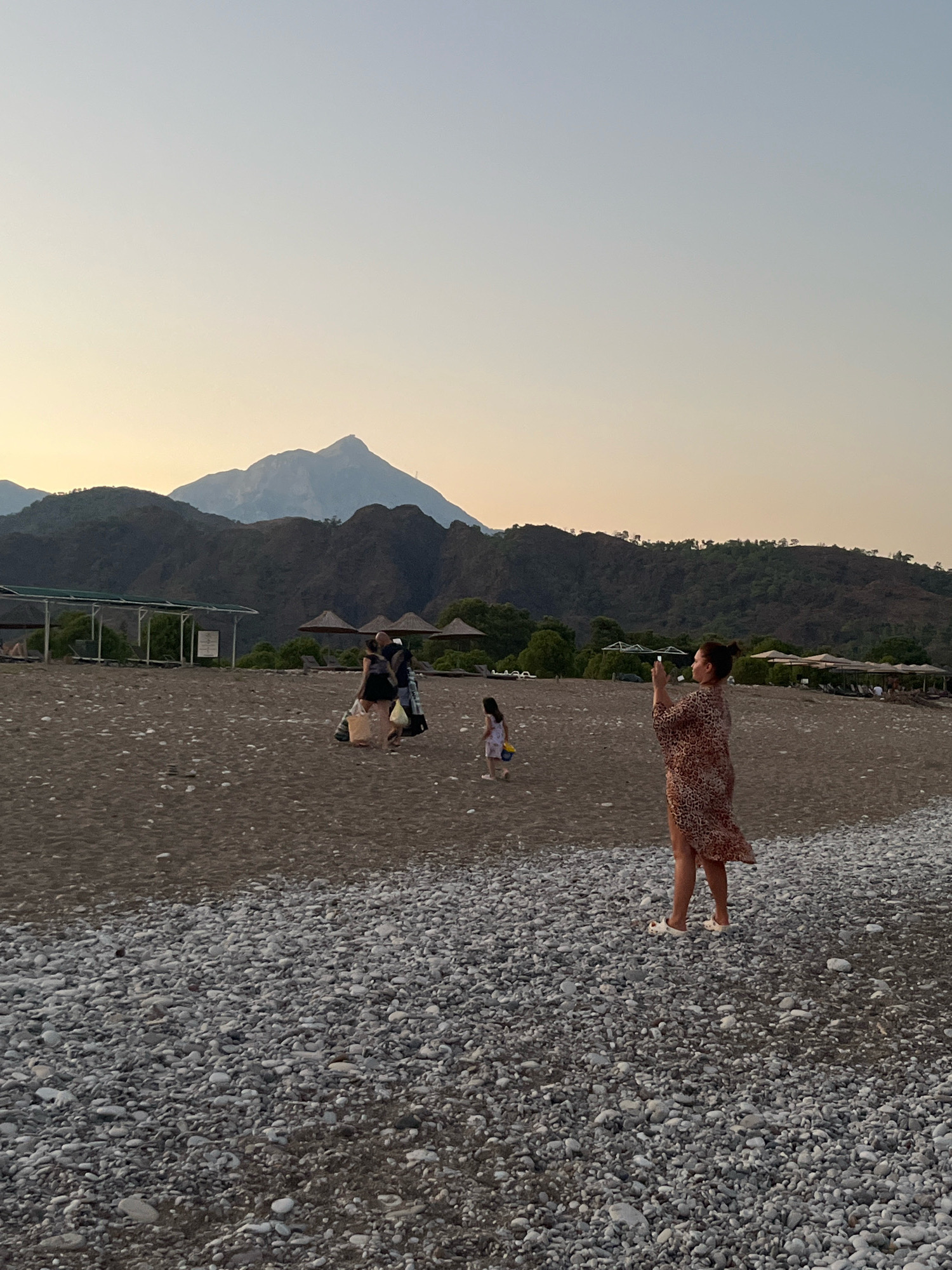 In the city of Kemer, Turkey, a woman with her hair tied up in a bun, wearing a leopard-print dress and white flats, stands on a pebble beach and holds up a phone to take a photograph. The beach is lined with beach umbrellas and loungers in the background. In the middle ground, a group of three people walk together, and a young child in a white dress walks alone. A mountain range is visible in the distance, with a prominent peak dominating the horizon. The sky above is a soft gradient of pale blue and yellow, indicating either early morning or late evening. The scene captures a serene moment on the Turkish coast.