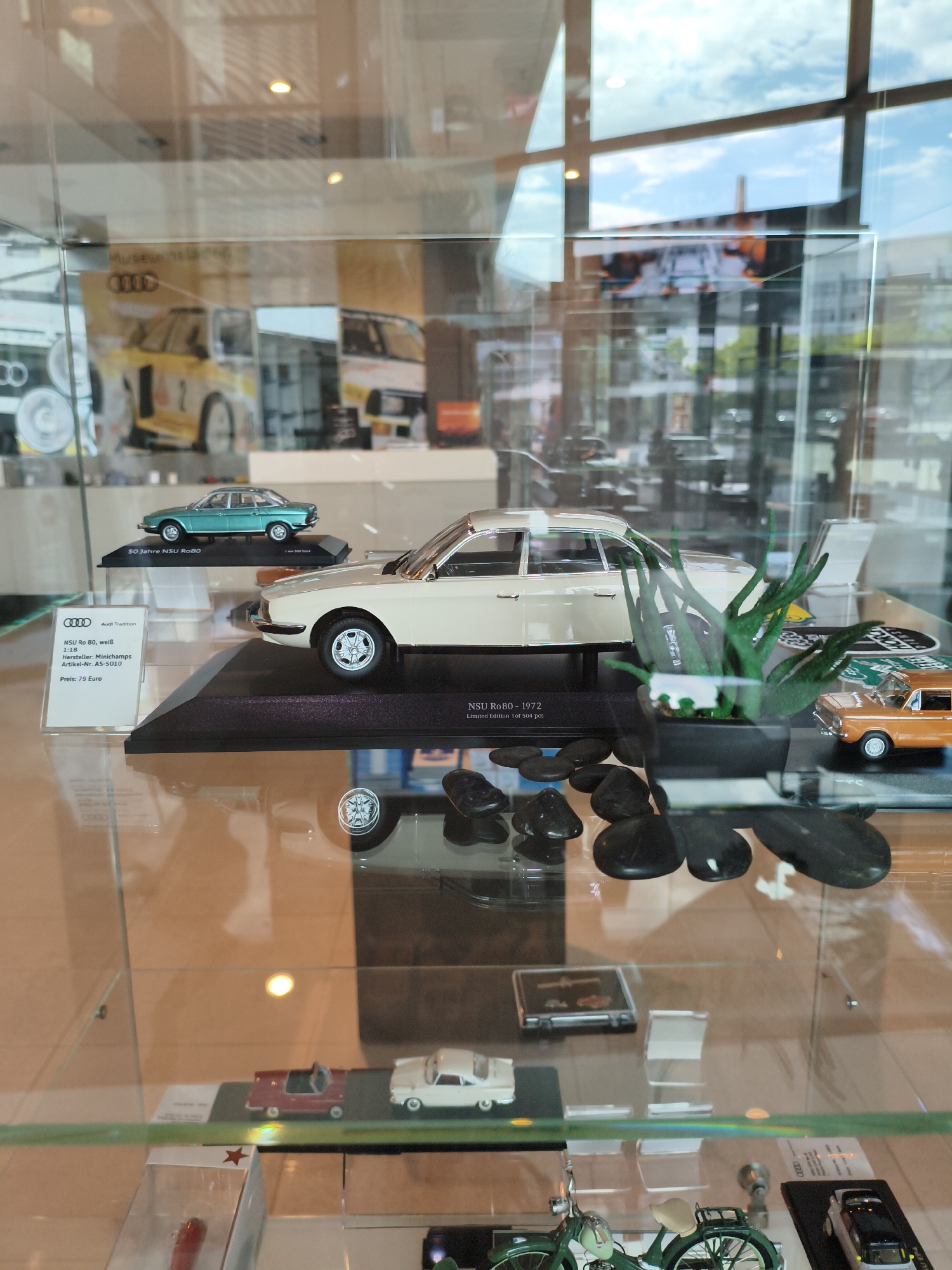The image shows a display case in the Audi Tradition Museum in Ingolstadt, Germany. The display case contains several miniature model cars and a plant. In the foreground, there is a beige model car on a black display stand with a label that reads "NSU Ro 80 - 1972 Limited Edition 1 of 504 pcs". To the left of this model is a small informational card with an Audi logo and text that reads "Audi Tradition", "NSU Ro 80, weiß", "1:18", "Hersteller: MiniChamps", "Artikel-Nr. AS-5010", and "Preis: 79 Euro". To the right of the beige model car, there is an orange model car and a small green plant in a black pot. In the background, there are reflections of the museum environment, including other displays, lighting fixtures, and windows showing the sky and greenery outside. Further in the background, there are other model cars on display, including a light blue car and a yellow car with the number "1" on it. The display case is made of glass, and the lighting inside creates reflections that obscure some of the details.