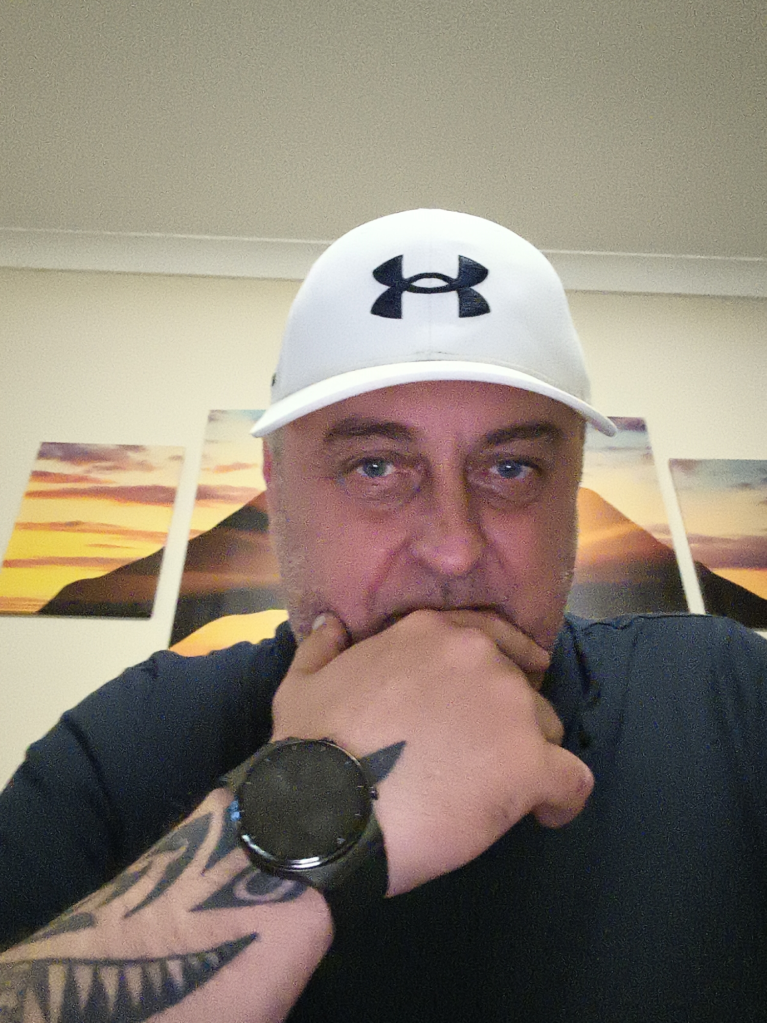 A close-up, slightly low-angle image features a man indoors in Huntingdonshire, United Kingdom. The man, appearing middle-aged, has blue eyes and looks directly at the camera. He wears a white baseball cap with a prominent black "Under Armour" logo. His right hand is raised, with his thumb resting on his chin and fingers covering his mouth, conveying a thoughtful or pensive demeanor. He is dressed in a dark-colored long-sleeved top. On his left wrist, he wears a dark-faced smartwatch with a black strap, and a striking black tribal-style tattoo with sharp, angular patterns covers his left forearm. The background reveals a light-colored wall adorned with three rectangular canvas prints arranged horizontally. These artworks depict a vibrant sunset or sunrise over silhouetted landscapes, characterized by warm yellow, orange, and purple tones. A plain, light-colored ceiling is visible at the top of the frame. The indoor lighting appears artificial.