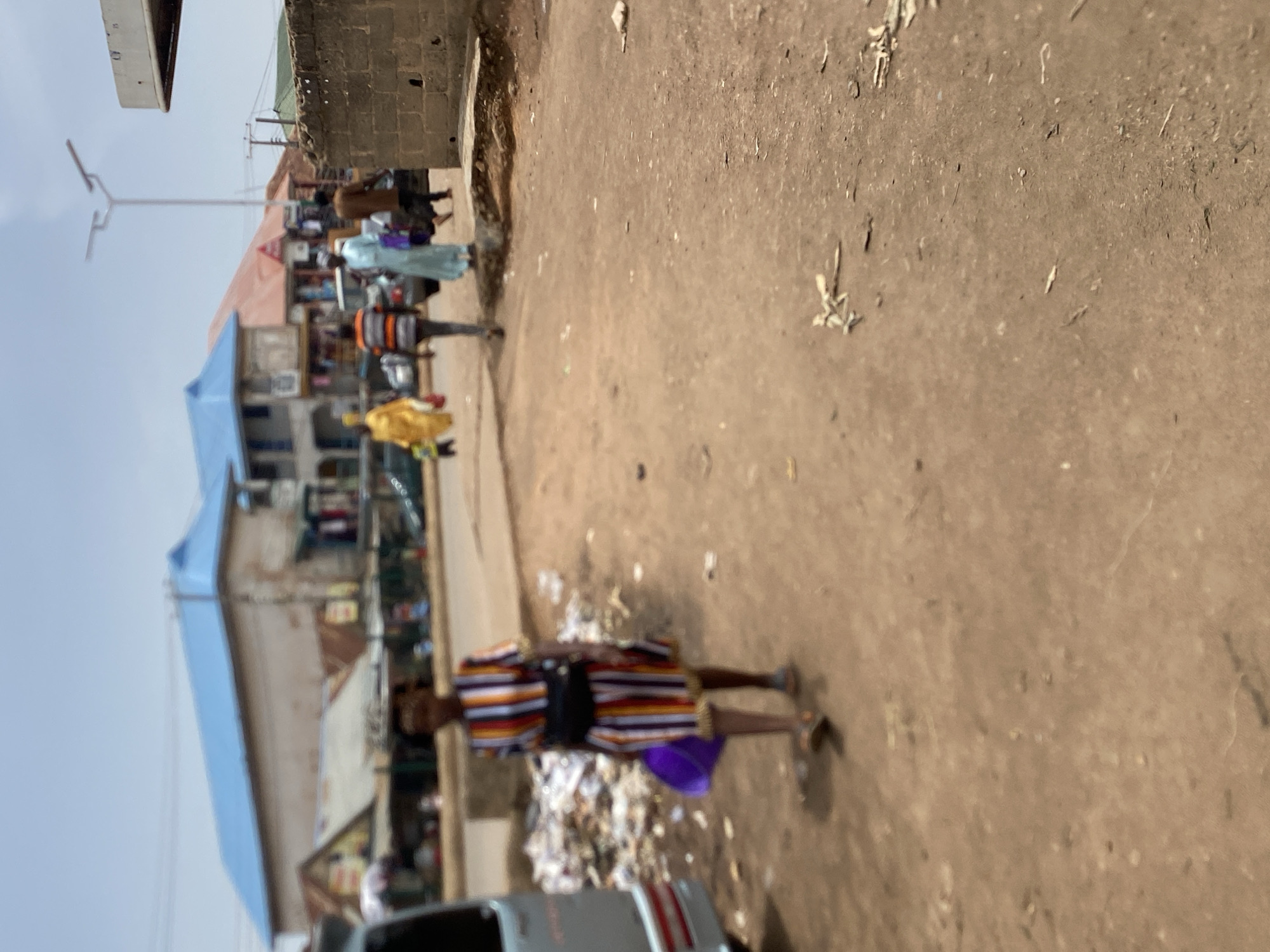 The image depicts a street in Bukuru, Nigeria, during the daytime. The ground is a mix of dirt and pavement. Litter is scattered around. Several people are walking in different directions. One person is wearing a striped outfit. Buildings are visible on the left side of the frame, with blue roofing. A car is partially visible. A street light is in the background.