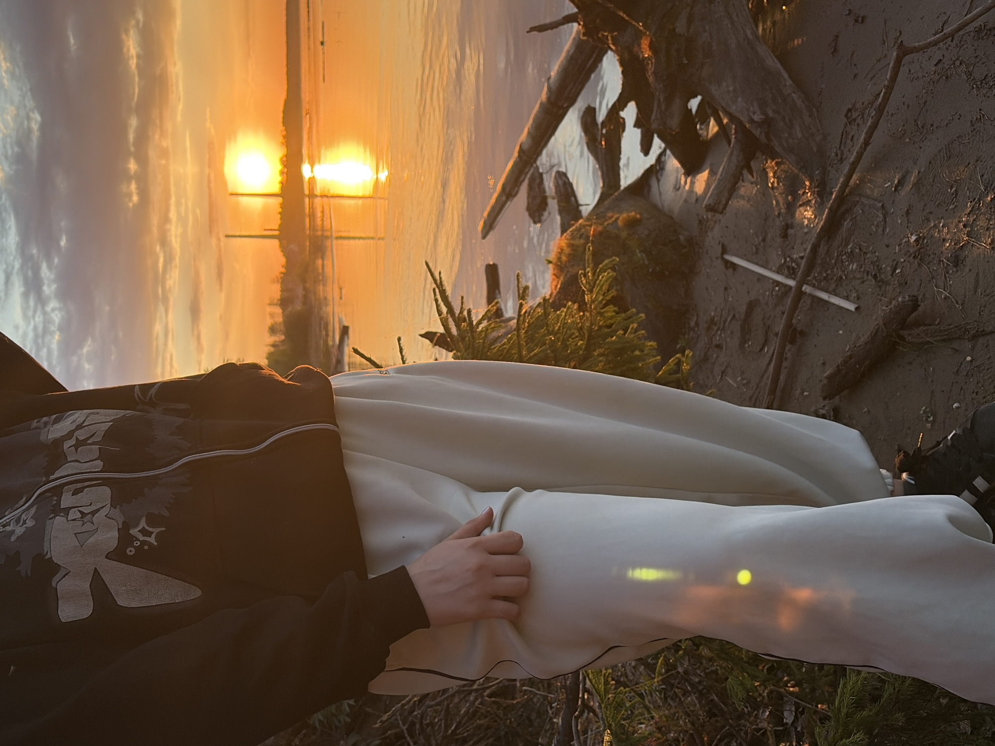 A horizontal photo shows a person lying near a shore. The person is wearing a dark zip-up hoodie with a light-colored design on the front left, and white pants. The person’s right hand is resting on their left thigh.
In the background is water reflecting a bright sunset. Trees and some vegetation are also visible. On the shore, there is driftwood and sand.
Based on the sunset over water, and the driftwood, this may be the shore of the Kama River, in Chaikovsky, Perm Krai, Russia.