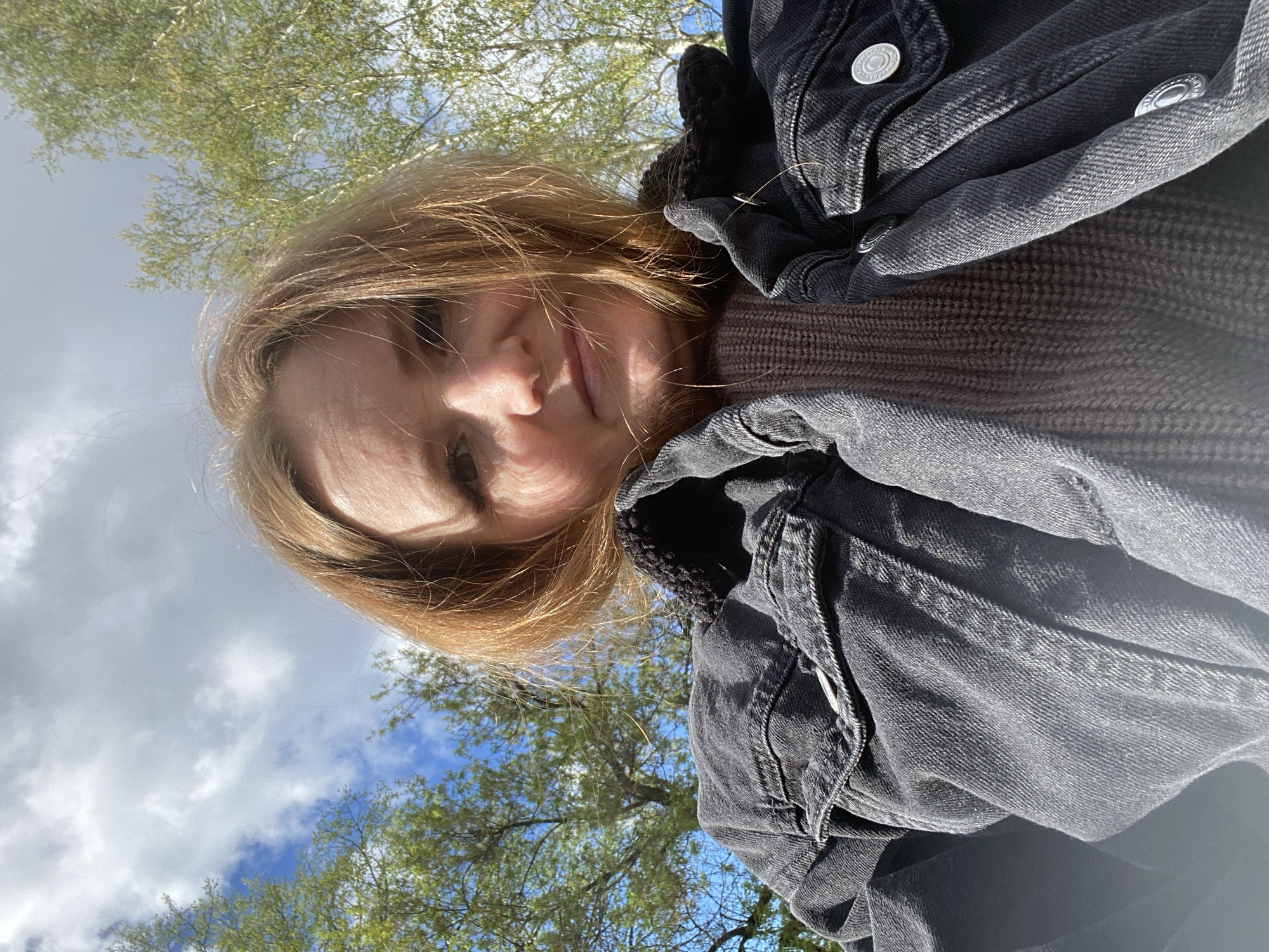 A selfie photograph shows a fair-skinned person with light brown hair. The person is wearing a dark grey denim jacket over a brown sweater. The background consists of blue sky with white clouds and tree branches with green leaves. The location is outdoors, and based on the description, it is possibly in the city of Chaikovsky, Russia. No specific activities or interactions are apparent.