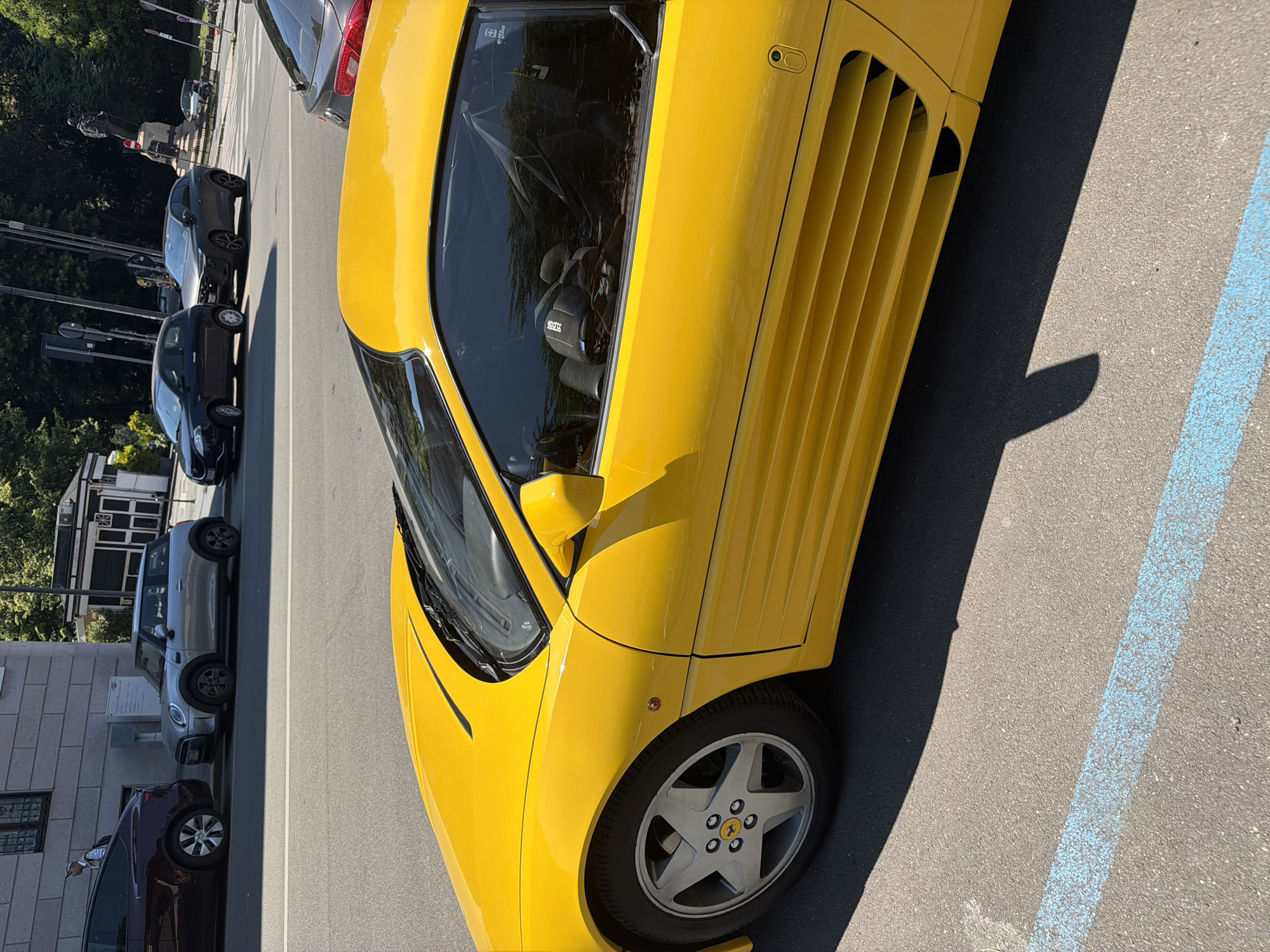 A bright yellow Ferrari sports car is parked in a designated parking spot marked with blue paint on asphalt. Several other vehicles, including cars in shades of gray, black, and dark maroon, are parked along the street. In the background, there's a building and trees, suggesting an urban environment. People are visible on the sidewalk near a street crossing with lamp posts. Based on the style of the buildings and overall aesthetics, this scene is located in the city of Milan, Italy.