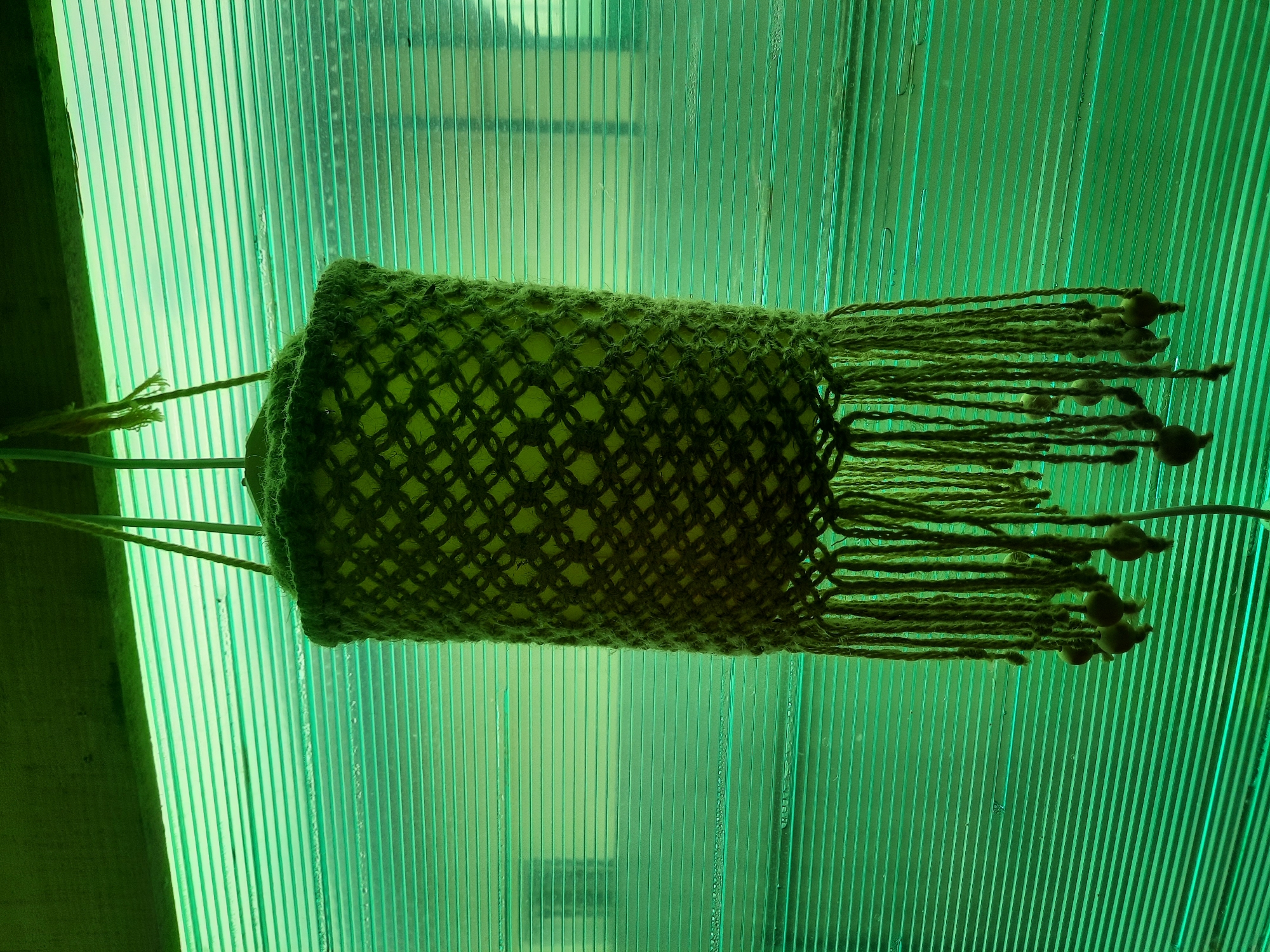 A macrame plant hanger with a cylindrical woven outer shell and a lighter green inner liner hangs against a translucent green corrugated plastic wall. The hanger is suspended by three strands of jute twine. The outer shell has a diamond-shaped pattern. The bottom of the hanger features a fringe of jute cords, some of which are adorned with small, dark wooden beads. The overall color palette is dominated by shades of green. The background wall suggests an indoor or covered outdoor space, possibly a greenhouse or porch. The location is stated to be Bolshoye Sorokino, Russia.