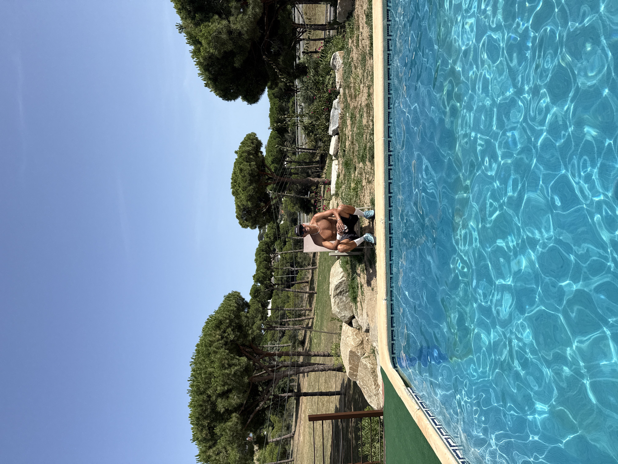 A young man, shirtless and wearing black shorts and a baseball cap, squats on a lounge chair beside a swimming pool. The pool water is a vibrant blue, reflecting the bright sunlight. Behind the pool, a grassy slope is dotted with rocks and mature pine trees. In the background, a highway can be seen, suggesting the location is near Sant Vicenç de Montalt, Spain, a town known for its proximity to coastal roads and natural landscapes. The sky is clear and a deep blue.
