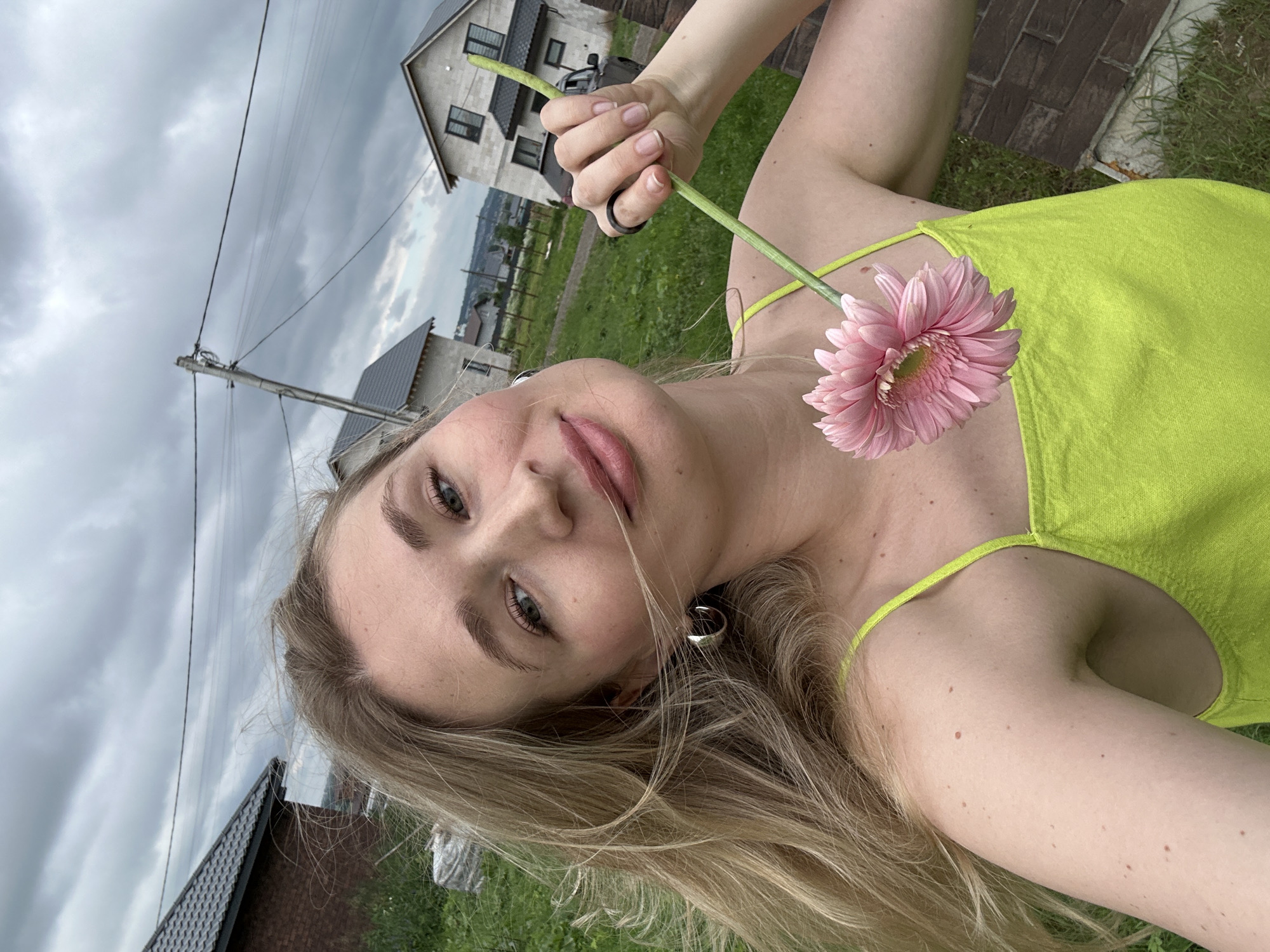 A young woman with blonde hair, wearing a lime green top, is holding a pink Gerbera daisy in her right hand. She is in a grassy outdoor area with several houses visible in the background. The sky is overcast. The location is the city of Mokino, Russia.