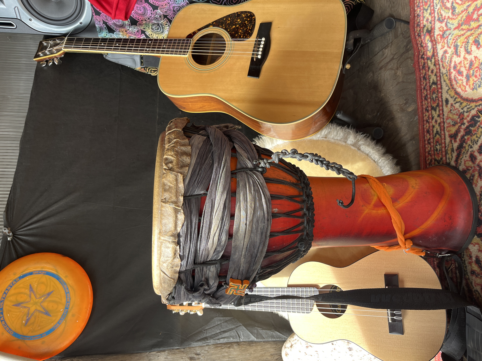 An acoustic guitar, ukulele, djembe drum, and frisbee are arranged in a cluttered indoor setting. The acoustic guitar, a light wood color with a tortoiseshell pickguard, leans against a black backdrop. To its left, a ukulele with a black strap is visible. In the foreground, a vibrant red djembe drum is wrapped in dark grey fabric and adorned with an orange sash. A frisbee, orange with blue accents and a star design, hangs on the black backdrop above the drum. The scene is lit by natural light, casting shadows and highlighting the textures of the instruments and surrounding objects. The floor is covered with a patterned rug.