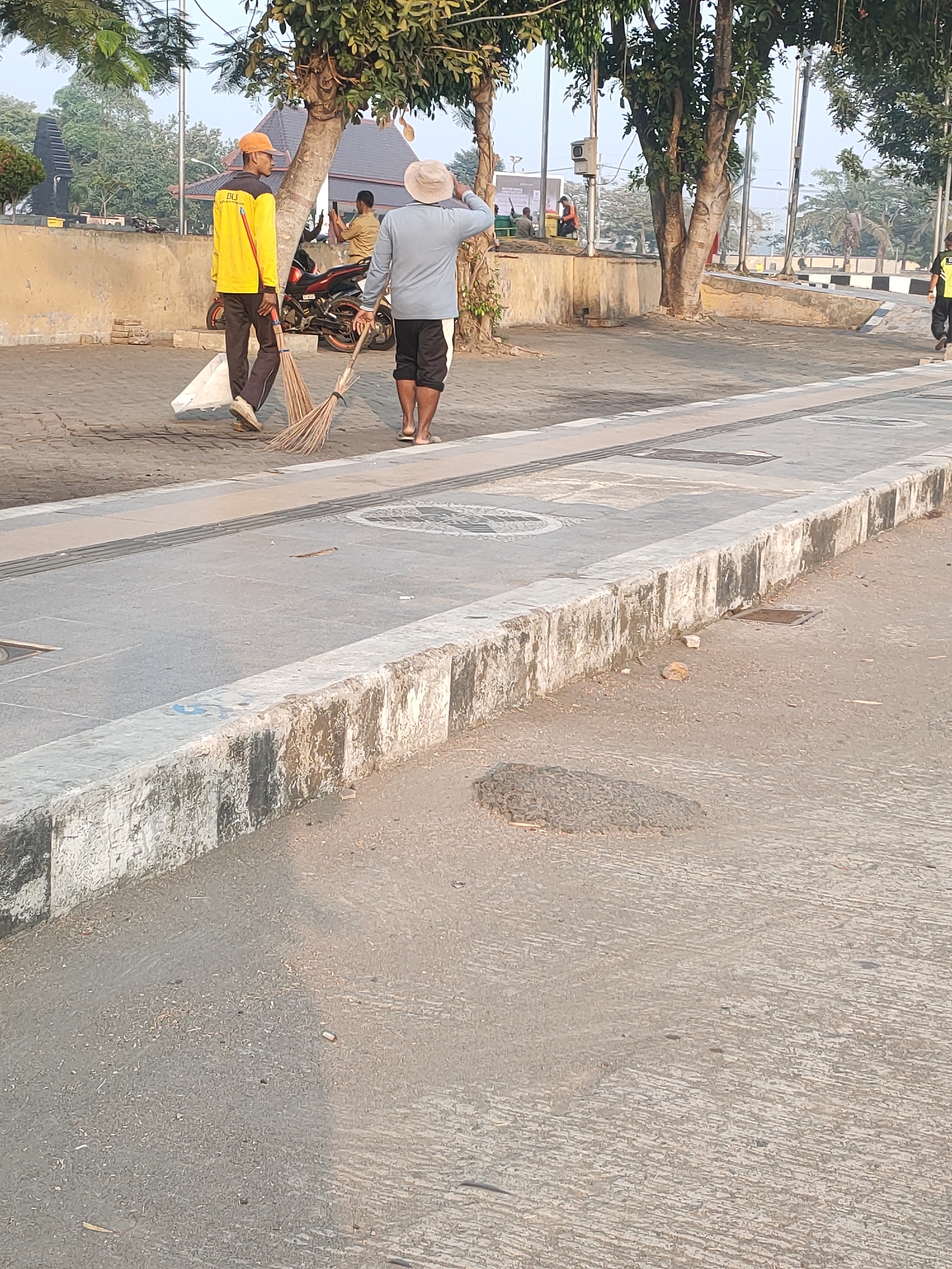 Two male sanitation workers are depicted in the image, actively sweeping a paved pathway. The worker on the left wears a bright yellow long-sleeved shirt with dark pants and an orange cap, holding a broom and a white bag. The worker on the right is dressed in a light blue long-sleeved shirt, dark shorts with white trim, and a light-colored hat, also holding a broom. They appear to be engaged in their duties in an outdoor urban setting. In the background, a motorcycle is parked, and several trees with lush foliage are visible, suggesting a public park or street area. The ground is a mix of pavement and dirt. This scene is located in the city of Kanigoro, Indonesia, likely a public area such as a street or park, given the presence of sanitation workers and urban elements.