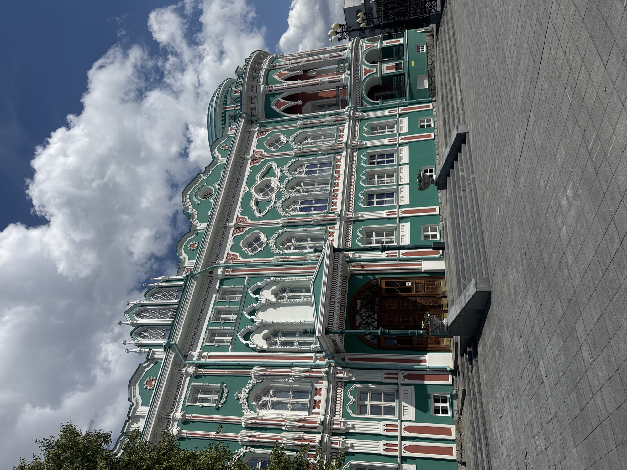 This image captures the Sevastyanov House, a historic mansion located in Yekaterinburg, Russia. The building is a prominent example of Gothic Revival architecture, characterized by its vibrant green facade adorned with white and red decorative elements. Ornate window frames, arched doorways, and decorative spires contribute to its distinctive appearance. The mansion stands on a wide, paved plaza with large stone steps leading up to its entrance. The sky above is a clear blue, punctuated by dramatic white clouds, indicating a bright, sunny day. A small tree with green foliage is visible on the left side of the frame. The overall scene conveys a sense of grandeur and historical significance.