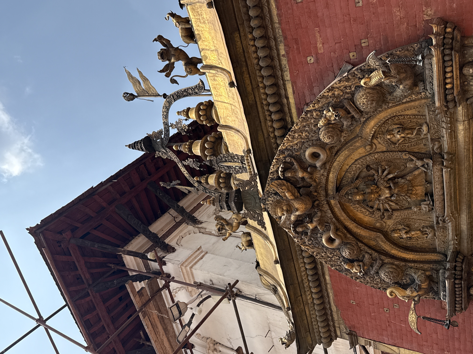 The image captures a low-angle view of the exterior of a temple building in Bhaktapur, Nepal. The building features intricate wood carvings and embellishments. The red brick wall is visible behind the decorative wooden elements, adding a contrast to the golden hue of the sculptures. Above the red brickwork is a series of ornate gold carvings, including figures of lions and other mythical creatures. Below the gold carvings is a white wall that is partially covered with scaffolding. The background is a clear, blue sky.