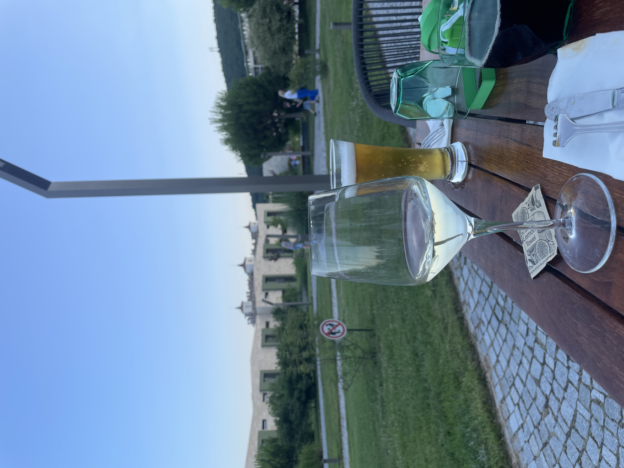 A table at an outdoor restaurant in Obzor, Bulgaria, features two glasses of drinks and some cutlery. In the foreground, a glass of white wine sits next to a beer. A dark liquid, likely a beverage, is in a glass to the right. A green glass is placed upside down on the table. Behind the table, a grassy area with cobblestone pathways and trees is visible. In the background, a building complex with unique architecture and a clear blue sky are observed. A sign with a red circle and a diagonal line through it, indicating a prohibition, is planted in the grass. Two individuals are walking in the distance.