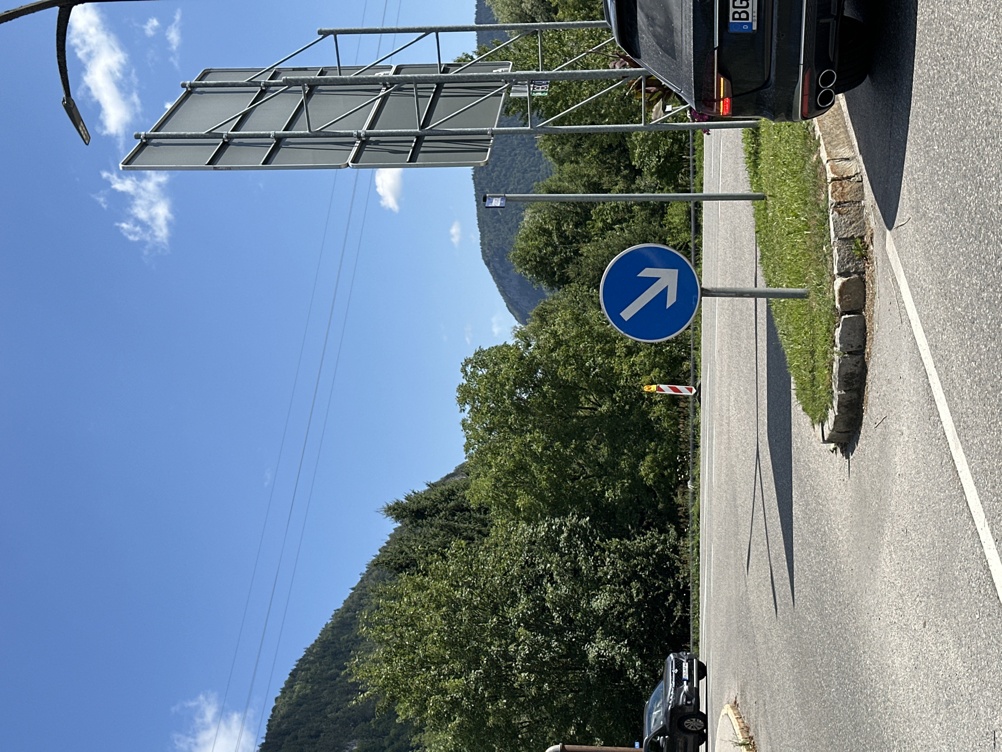 The image shows a road intersection in Berchtesgaden, Germany, on a sunny day. A black SUV is parked on the right side of the road, partially obscuring a large, gray advertising billboard supported by a metal structure. In the center of the image, a blue circular traffic sign with a white arrow pointing diagonally down and to the left is mounted on a pole. A black car is driving on the road to the left. The background features lush green trees and rolling hills under a clear blue sky with a few scattered clouds. The road is paved asphalt with white lane markings.