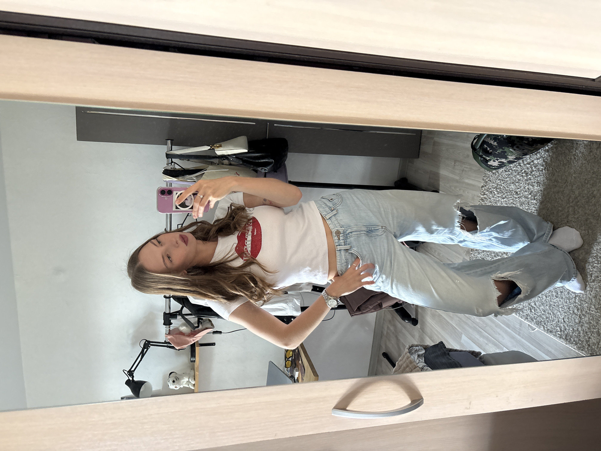 A young woman with long, light brown hair and fair skin is reflected in a mirror, taking a selfie. She is wearing a white t-shirt with a large red lip graphic on the front and light-wash, distressed denim jeans. She has a silver watch on her left wrist and a ring on her right index finger. The setting appears to be an indoor space, possibly a bedroom or dressing area, with a wardrobe visible in the background. In the reflection, there are shelves with various items, including bags, shoes, and a desk lamp. The floor is covered with a carpet and wooden planks. The lighting suggests it is daytime.