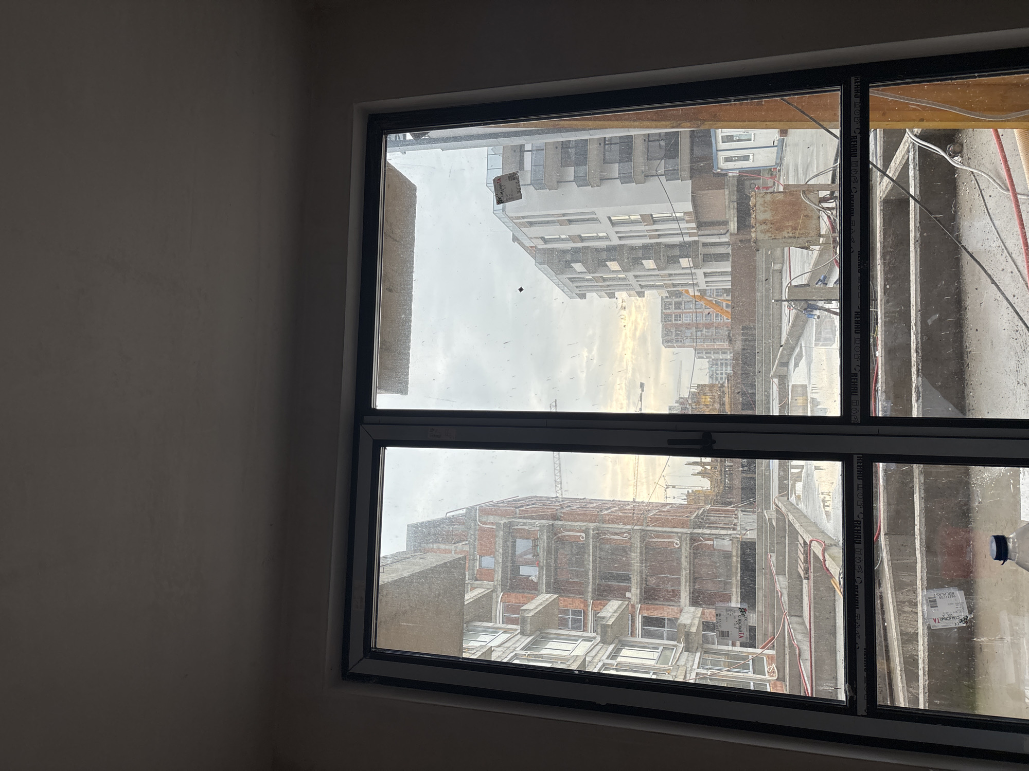 View from an interior space through a double window, displaying an outdoor scene. The scene reveals ongoing construction projects with unfinished buildings, scaffolding, and construction equipment, possibly cranes, in view. Cables and construction materials are visible near the window frame on the right. The urban environment is evident through the view of buildings. A sticker is visible on the window pane. The location is suggested to be Burgas, Bulgaria, based on the context. No people are visible in the scene. The lighting suggests an overcast day.