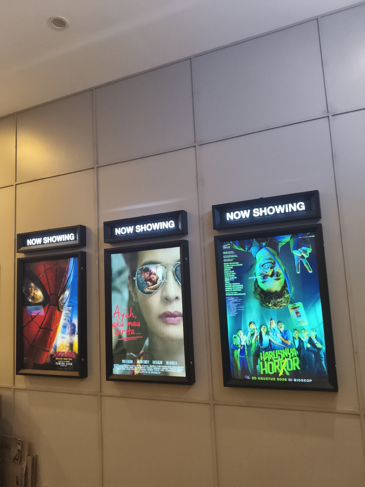 The image shows three movie posters on a wall in a cinema lobby. The wall is paneled with a light beige color. Above each poster is a lit sign displaying "NOW SHOWING" in white text against a black background.
The leftmost poster features a close-up of Spider-Man's mask, with the "SPIDER-MAN: GRAND NEW DAY" title and "COMING SOON" text visible.
The middle poster shows a close-up of a woman's face wearing sunglasses, with a reflection of a couple in the lenses. The text "Ayah, aku mau cerita..." is prominent.
The rightmost poster is for a horror movie titled "HARUSNYA HOROR" (Horror). It displays a group of people with expressions of fear and surprise, with a man upside down in the center. The release date "20 AGUSTUS 2026 DI BIOSKOP" (August 20, 2026 in Cinema) is at the bottom.
The lighting in the scene is a mix of warm yellow from overhead lights and the illuminated posters. To the far left, a stack of newspapers or magazines is visible. The overall environment suggests a modern cinema setting.