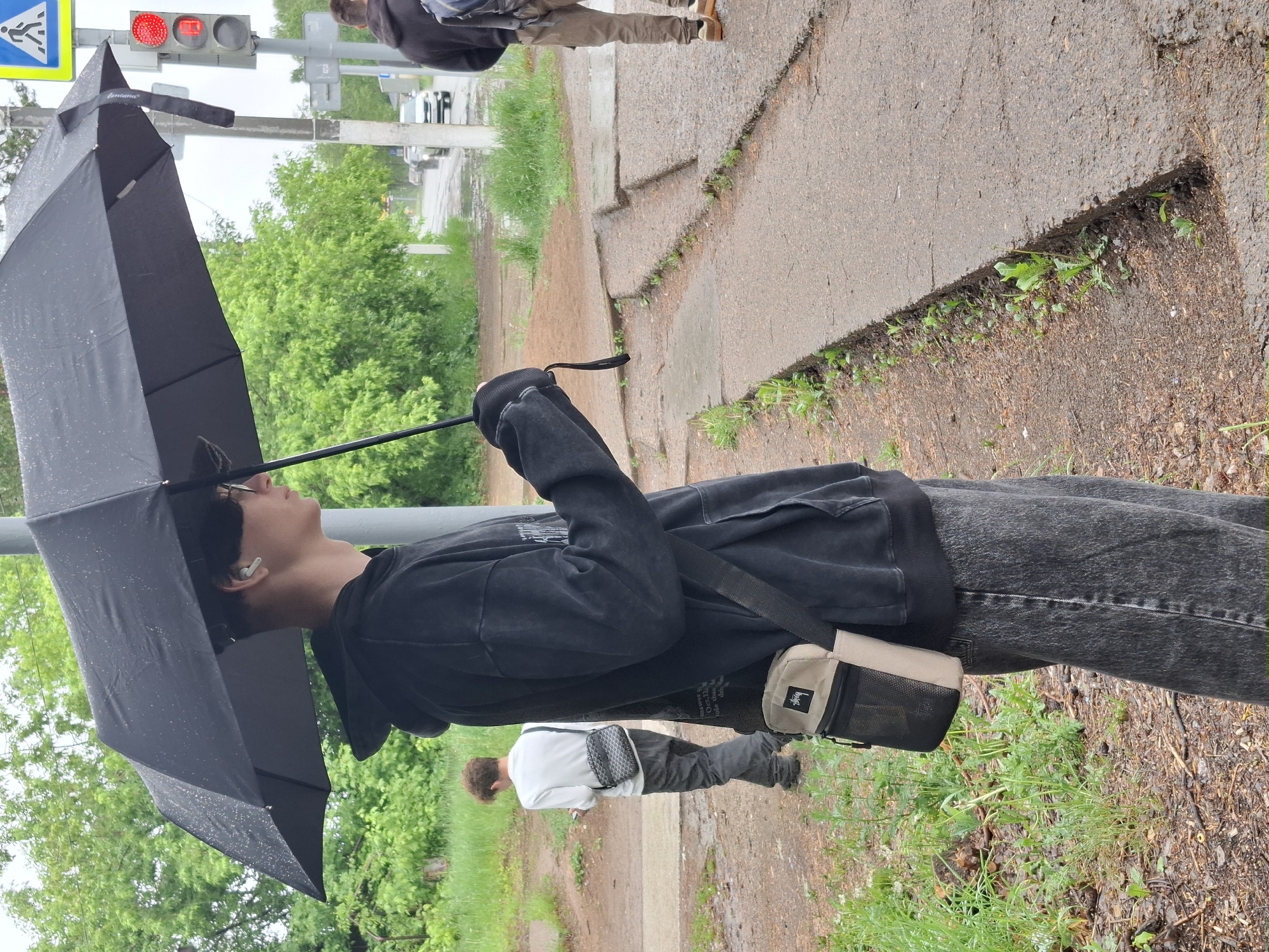 A young man in a black hoodie, grey jeans, and a crossbody bag walks down a sidewalk in the city of Chaikovsky, Russia, holding a black umbrella over his head. The traffic light next to him is red. Other people can be seen walking in the background. The environment includes trees, grass, sidewalks, and roads.