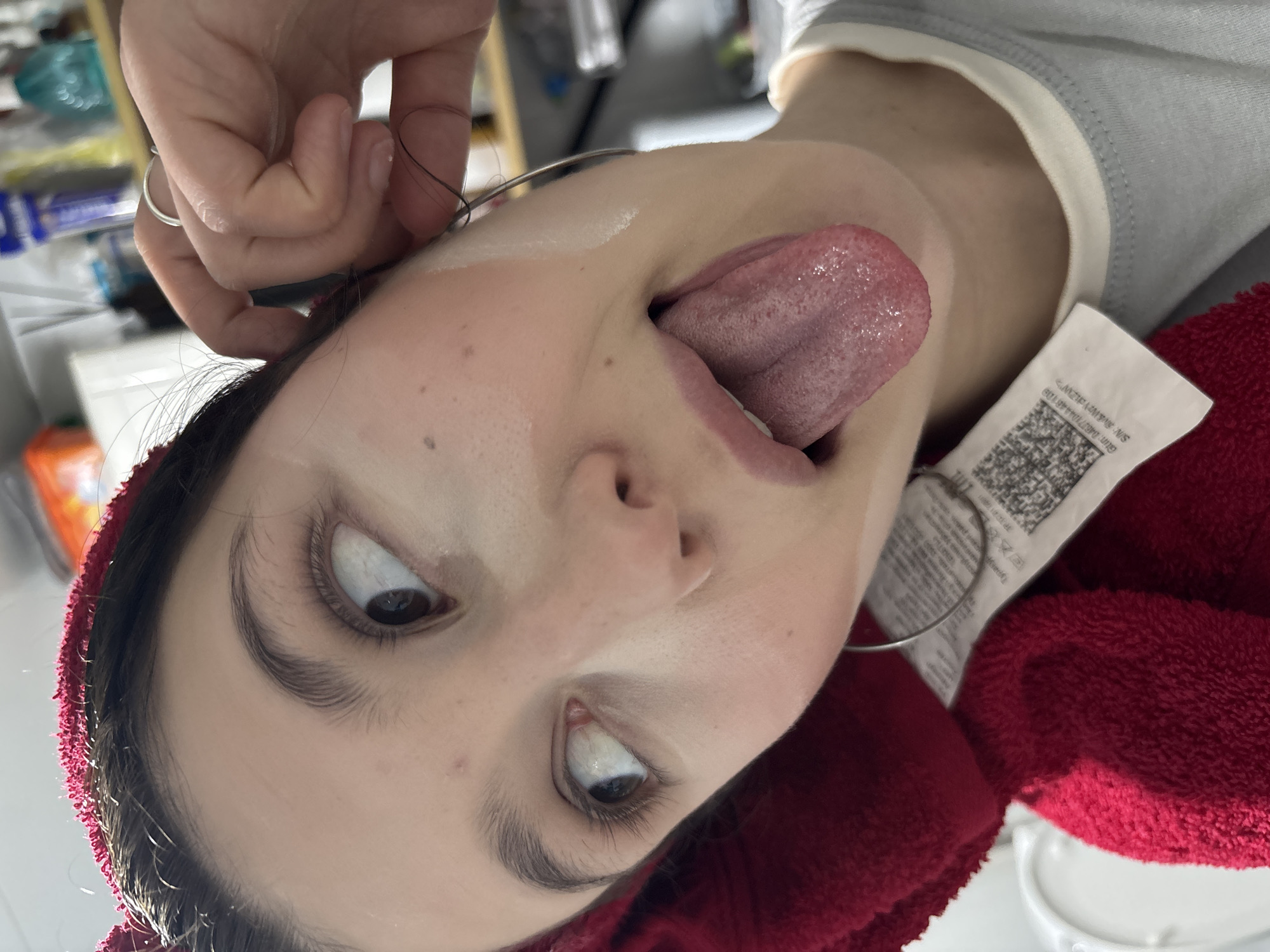 A woman is positioned upside down, sticking her tongue out. Her eyes are looking to the side. A hand is holding what appears to be a thin metal wire near her head. The subject is wearing a light gray top with a white collar, and a red towel is draped around her neck. A clothing tag is visible near her face. The background is blurry, showing what appears to be a kitchen or bathroom setting. The location Фоки, Russia, is not discernible from the image.