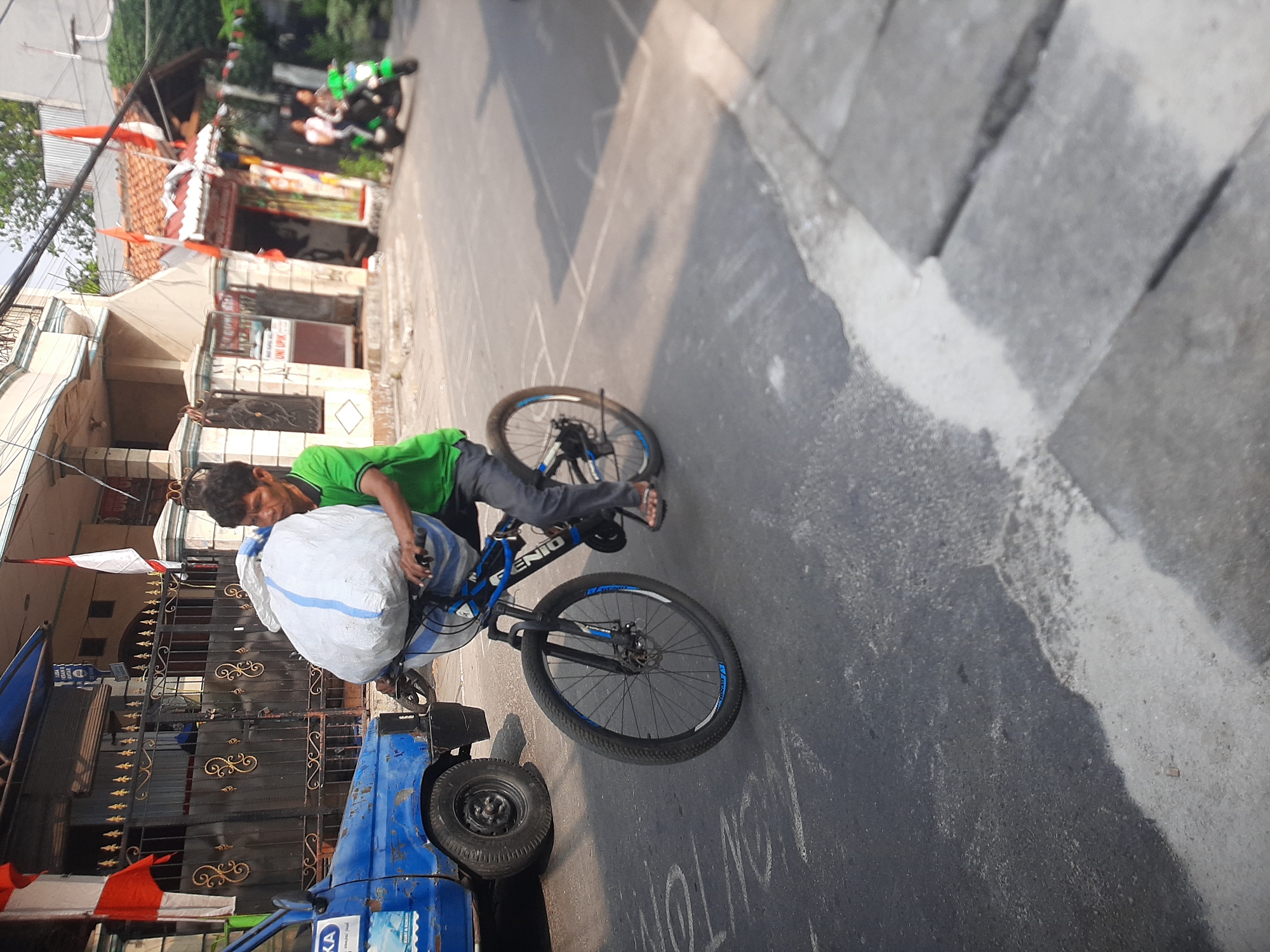 A medium shot captured on a sunny day in Central Jakarta, Indonesia shows a man riding a bicycle on a paved road. The man is wearing a green t-shirt and dark pants, and he is pedaling a blue and black bicycle with a large white bag secured to the handlebars. The bag appears to be filled with bulky items. To the left of the bicycle, a blue truck is parked on the side of the road. The background features a building with a dark metal gate, and flags of Indonesia are visible. In the distance, a green motorcycle carrying two people is also on the road. The scene appears to be a typical street in a residential or commercial area.