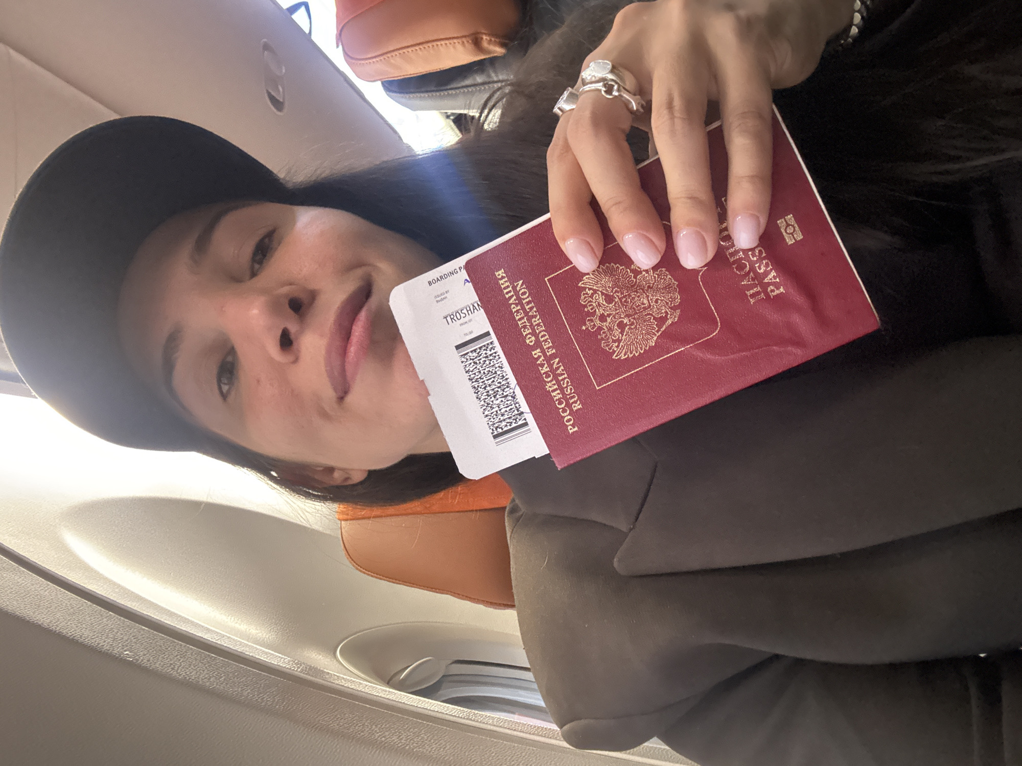 A woman is on an airplane, holding a boarding pass and what appears to be a Russian passport. The boarding pass includes the name "TROSHAN". The image does not provide any location details regarding the city of Khimki, Russia.