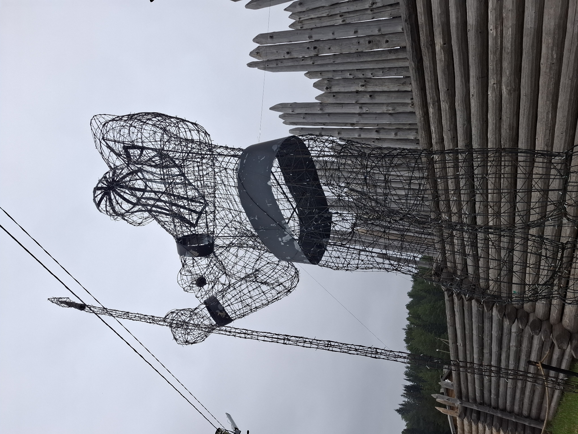A wire sculpture of a man stands in front of a wooden palisade in Adam, Russia. The sculpture depicts a figure with a spear. The palisade has both vertical log sections and angled, flat planks. The sky is overcast.
