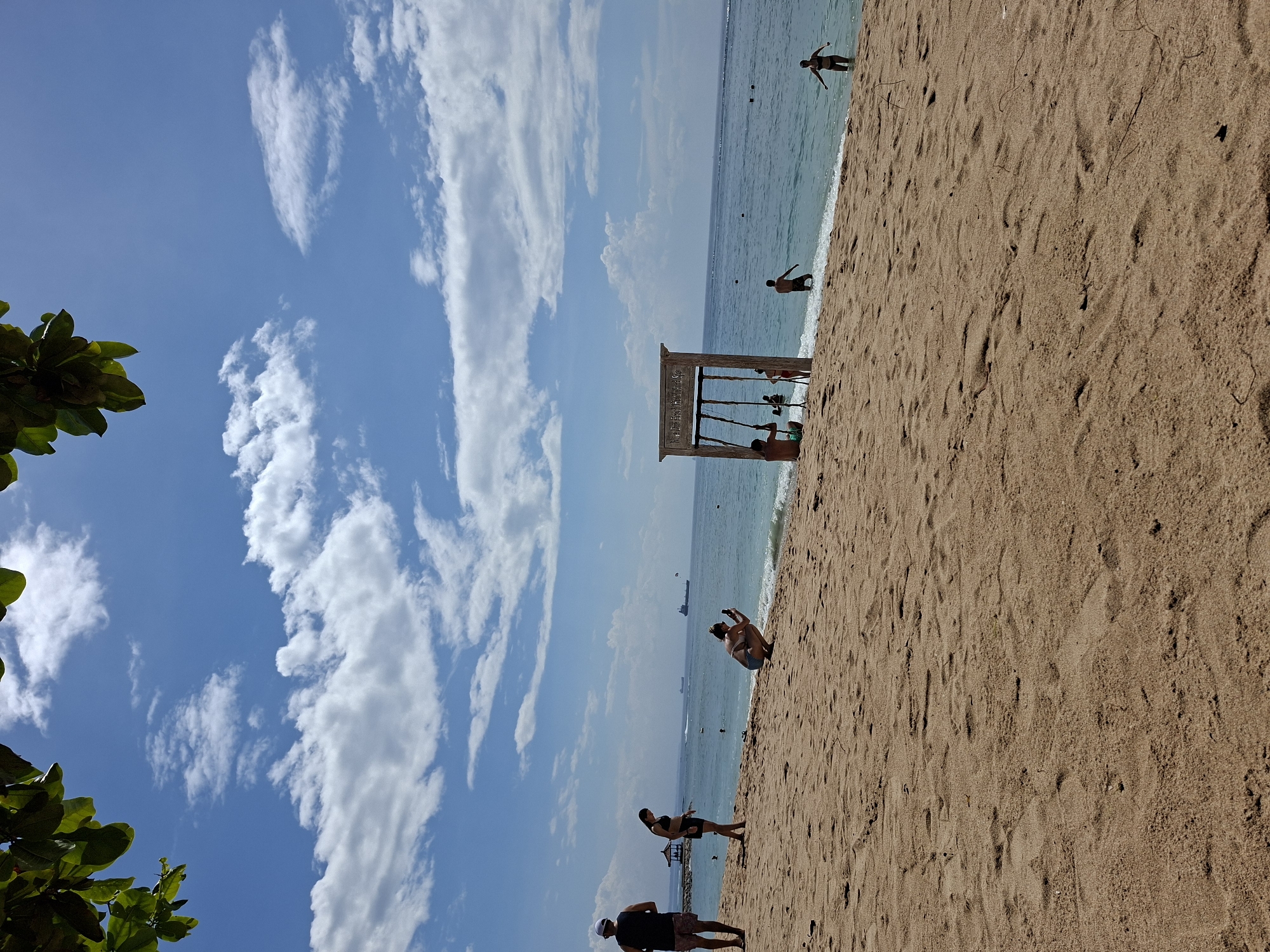 This is a bright daytime scene capturing a lively beach environment in Kuta Selatan, Indonesia, likely near the Nusa Dua area. The image features a clear blue sky filled with white, wispy clouds in the upper portion, transitioning to the calm, turquoise ocean and light brown sandy beach.
In the bottom-left corner, vibrant green leaves of a tree or bush frame the shot. The expansive sandy beach, stretching from the bottom-right across the midground, shows faint ripples and footprints. Several people are present on the sand: one individual in dark attire stands towards the bottom-right, another further up the beach in a black top, and a person in light swimwear is crouched, possibly taking a picture.
A distinctive wooden arch-like swing set is positioned at the water's edge, with some parts in the shallow water. Visible text on the structure reads "Nusa Dua Beach Hotel & Spa," serving as a clear location cue. Multiple individuals are seen around this swing set, with some seemingly engaging with the swings.
The serene ocean extends into the background, where several people are wading or swimming near the shore. Far on the horizon, distinct silhouettes of ships or boats are visible. The overall atmosphere suggests a sunny, pleasant day, ideal for beach activities and relaxation.