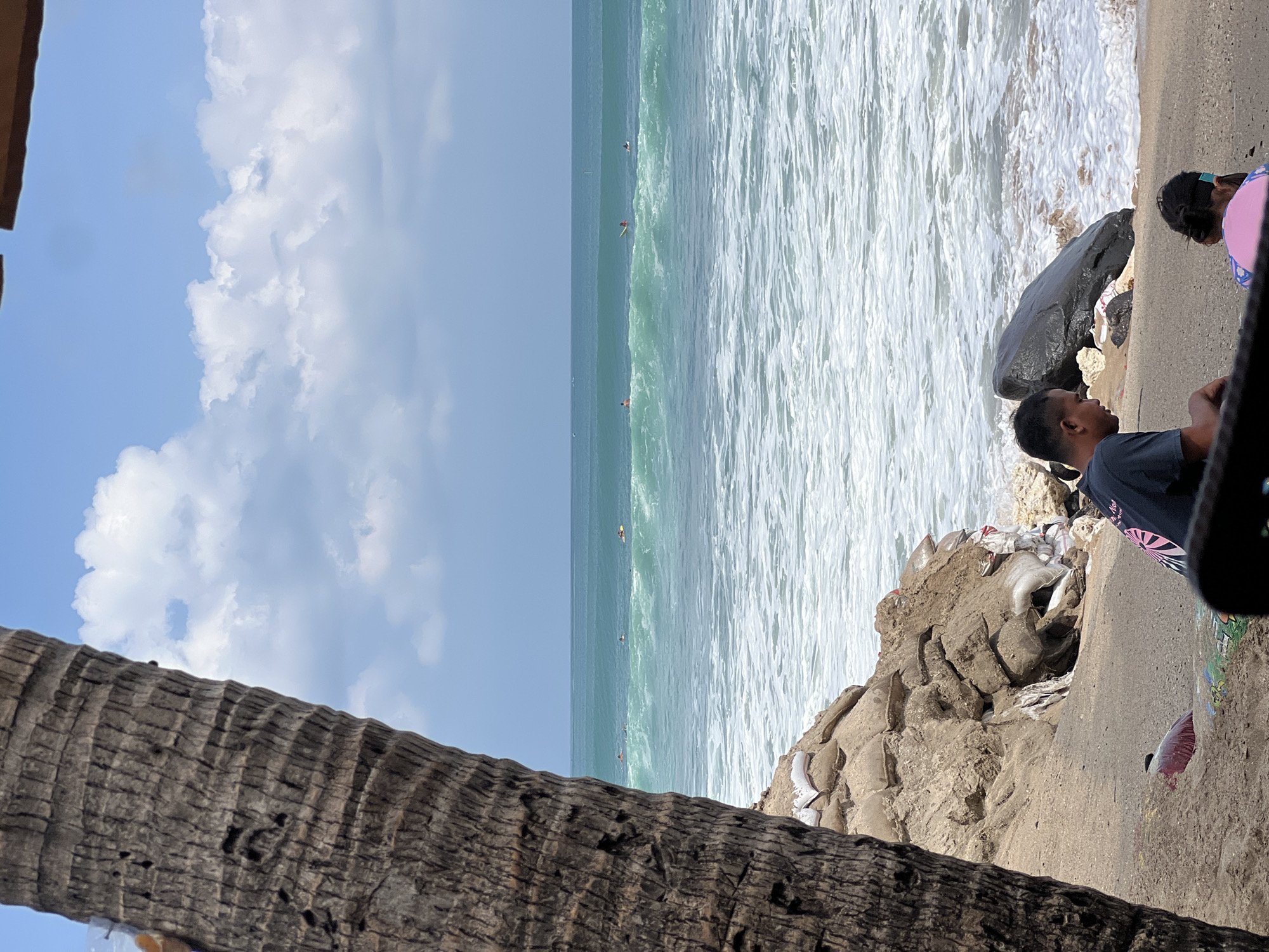 The image depicts a beach scene in Kuta, Indonesia, with a focus on the ocean and the shore. In the foreground, a large palm tree trunk occupies the left side of the frame, partially obscuring the view. On the sandy shore, a man and a child are visible, facing the ocean. The man is wearing a dark t-shirt, and the child is wearing a pink garment. A large dark rock is situated near the water's edge. The ocean exhibits turquoise waves, with several surfers scattered across the water. The sky is partly cloudy with a clear blue expanse. The shoreline appears to be reinforced with sandbags, suggesting erosion control measures. The overall scene captures a typical day at a popular Indonesian surf spot.