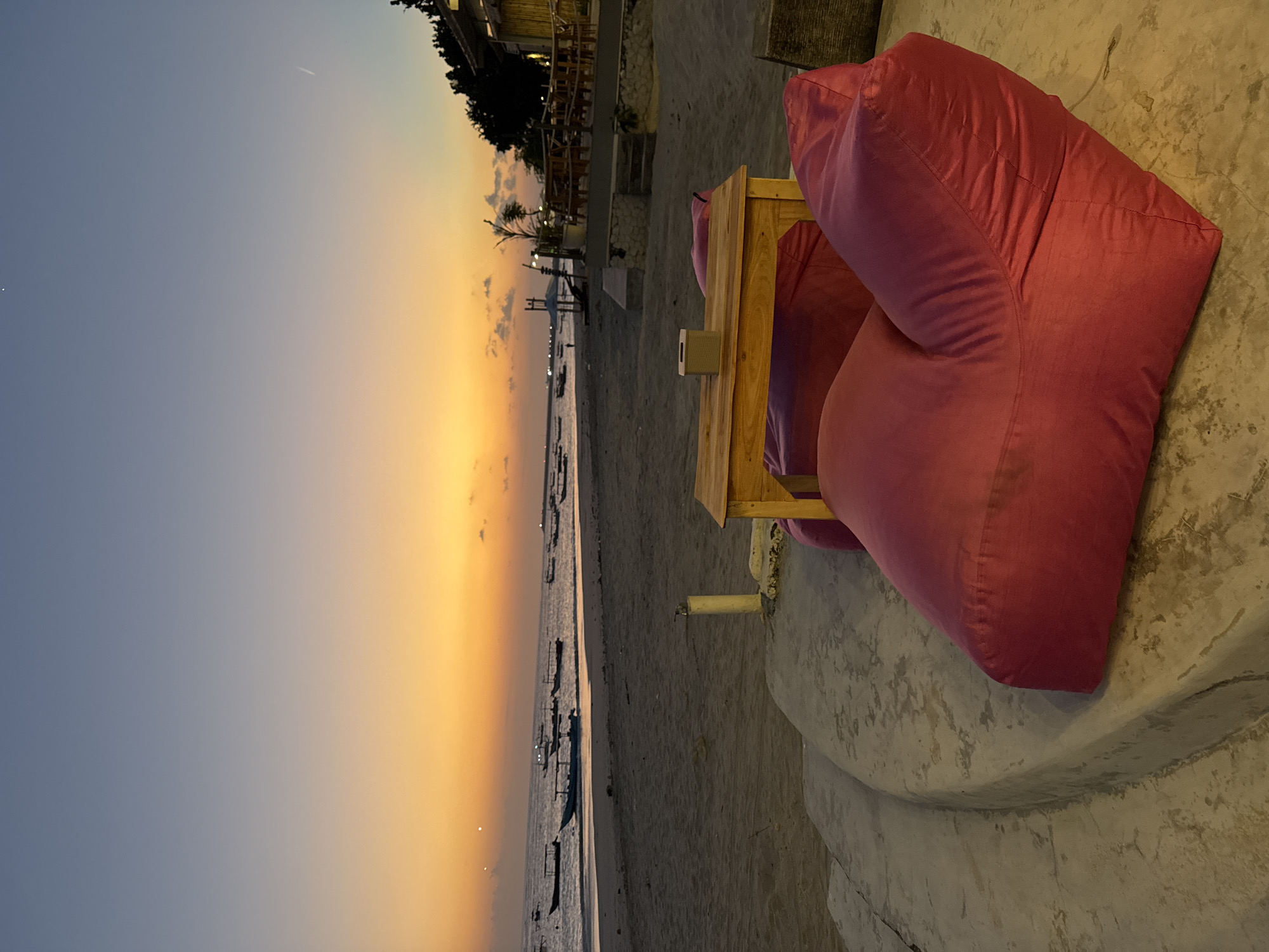 A twilight scene at the beach in Kuta, Indonesia, features a sandy shore with calm ocean waters reflecting the orange and purple hues of the sunset. Several traditional Indonesian fishing boats are anchored offshore. In the foreground, two large, plush pink beanbag chairs are positioned on a curved concrete platform, facing the ocean. A low wooden table sits between the beanbags, topped with a small, square tissue box. To the right of the beanbags, a raised wooden deck with multiple tables and chairs is visible, suggesting a beachfront cafe or restaurant. The environment appears serene and inviting, capturing the peaceful ambiance of a coastal evening.