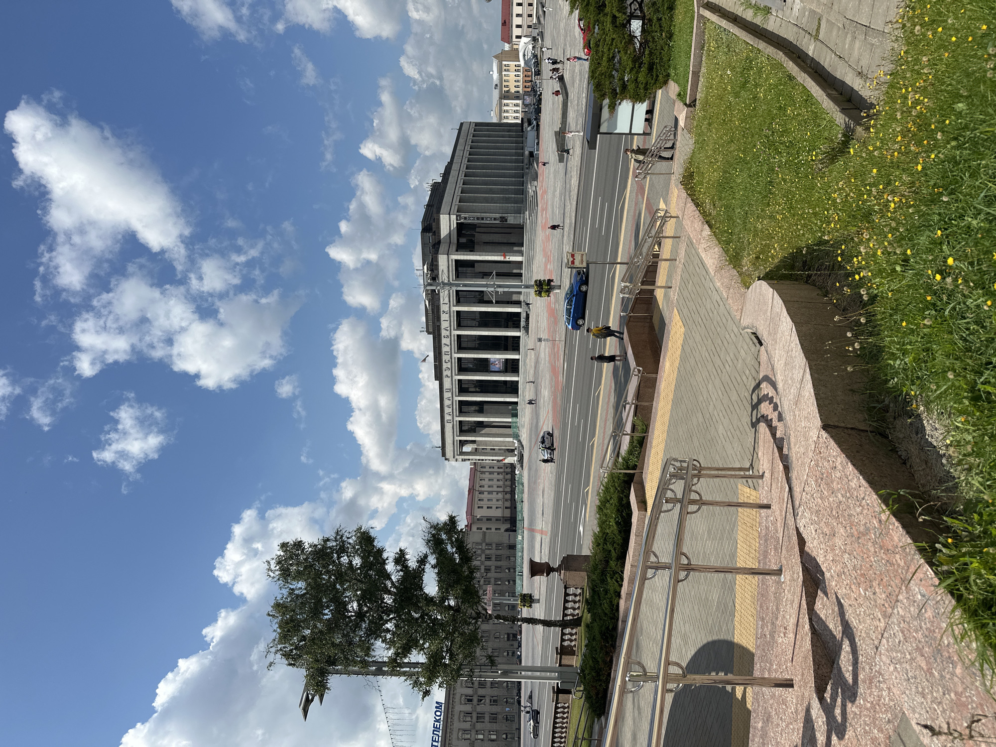 The image shows a wide outdoor plaza in Minsk, Belarus, dominated by a large neoclassical building with a prominent colonnade. The building has "ПАЛАЦ РЭСПУБЛIКI" (Palace of the Republic) written in large letters across its facade. The plaza is paved with light-colored stones and features several lanes of traffic. A lone tree stands in the middle of the plaza near the foreground. Several people are visible walking in the plaza and near the building. A blue car is driving across the plaza, and other cars are parked or moving in the background. The sky is bright blue with scattered white clouds. In the foreground, there are stairs leading down from a raised viewpoint, with metal handrails and stone steps. A patch of green grass with small yellow flowers is situated next to the stairs. The overall scene depicts a sunny day in a public urban space.