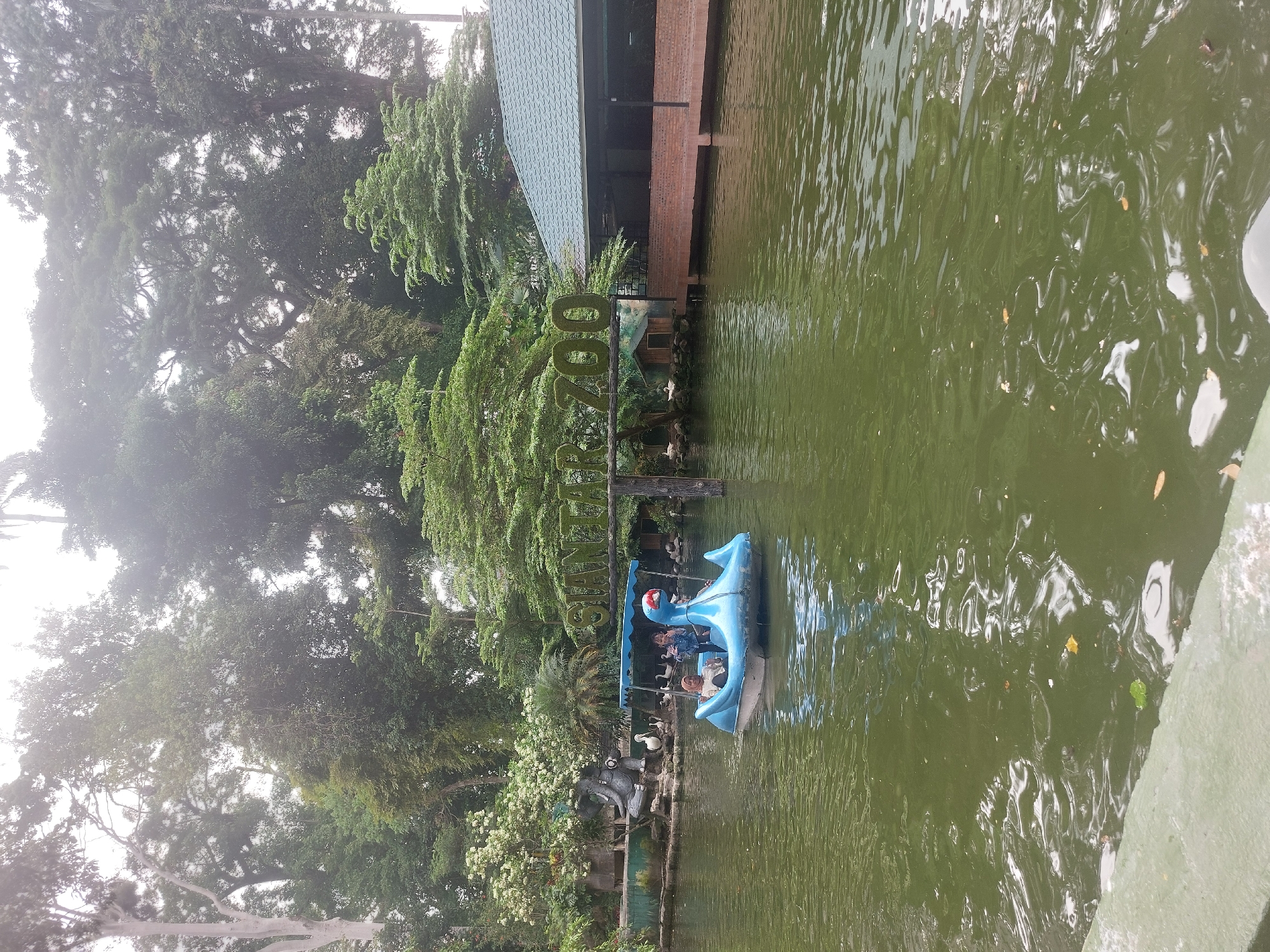 In the city of Pematang Siantar, Indonesia, a blue swan-shaped pedal boat carrying two young girls is floating on a body of water. The girl in the front is wearing a denim jacket and the girl behind her is wearing a hijab and a light-colored top. The water is green and rippling. In the background, there is a sign made of mossy letters that reads "SIANTAR ZOO." To the right of the sign, a building with a green roof and brick walls is visible. To the left of the sign, there is a stone sculpture of an elephant and several white birds. The scene is surrounded by lush green trees, creating a natural, park-like atmosphere. The sky is overcast, suggesting a cloudy day.