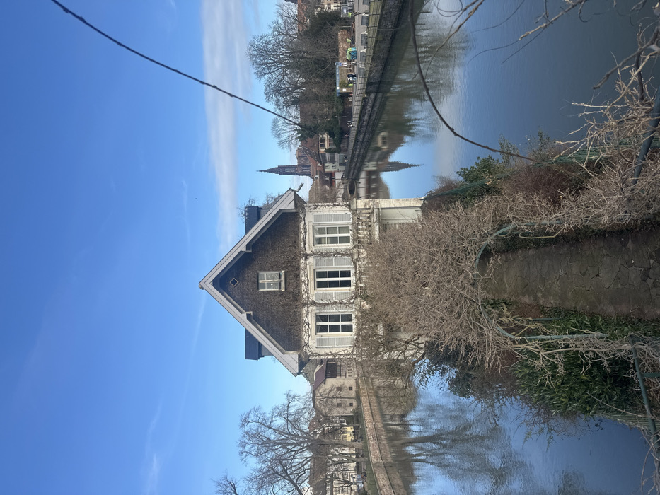 The image depicts a canal scene in the city of Strasbourg, France, likely within or near the Petite France district, characterized by a prominent house on a narrow land strip. The central subject is a two-story house with a steep, dark grey gable roof, adorned with a small diamond-shaped decorative element near its peak. The exterior walls are light-colored, featuring multiple white-shuttered windows, and the facade is partially covered by leafless climbing vines.
The house stands directly adjacent to calm, reflective canal water on at least two sides. In the immediate foreground, a narrow stone pathway, composed of irregular flagstones, leads towards the house, bordered by leafless woody vines trained over several metal archways and dense evergreen bushes. A thin, dark, out-of-focus branch extends into the upper right foreground, partially obscuring the view.
To the left of the house, the canal continues, with distant buildings and leafless trees lining its bank. To the right, another section of the canal is visible, with a paved walkway along its far bank where several indistinct figures are present. Scattered leafless trees and a few structures, possibly outdoor art installations, are visible along this bank. In the far background, towering above the buildings and trees on the right, the distinct gothic spire of Strasbourg Cathedral is clearly visible, reflected in the canal water. The sky is a clear, bright blue with sparse, wispy white clouds. The overall lighting suggests a sunny day.