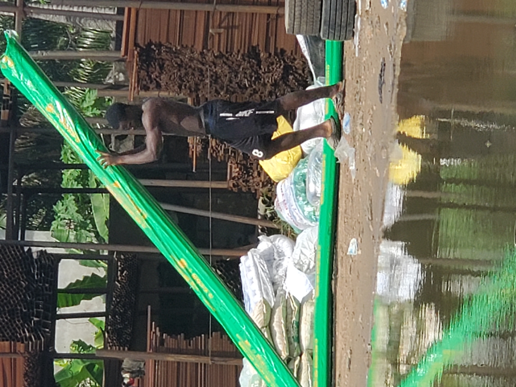 A shirtless man wearing black shorts stands on a green, flat-bottomed boat piled with sacks and packages. The boat is moored near a muddy shoreline with tire treads visible. Reflections of green and brown hues are visible in the nearby water. Behind the man and the boat is a partially visible structure made of wood and metal. Vegetation is visible behind the structure. The scene is at the city of Fegge, Nigeria.