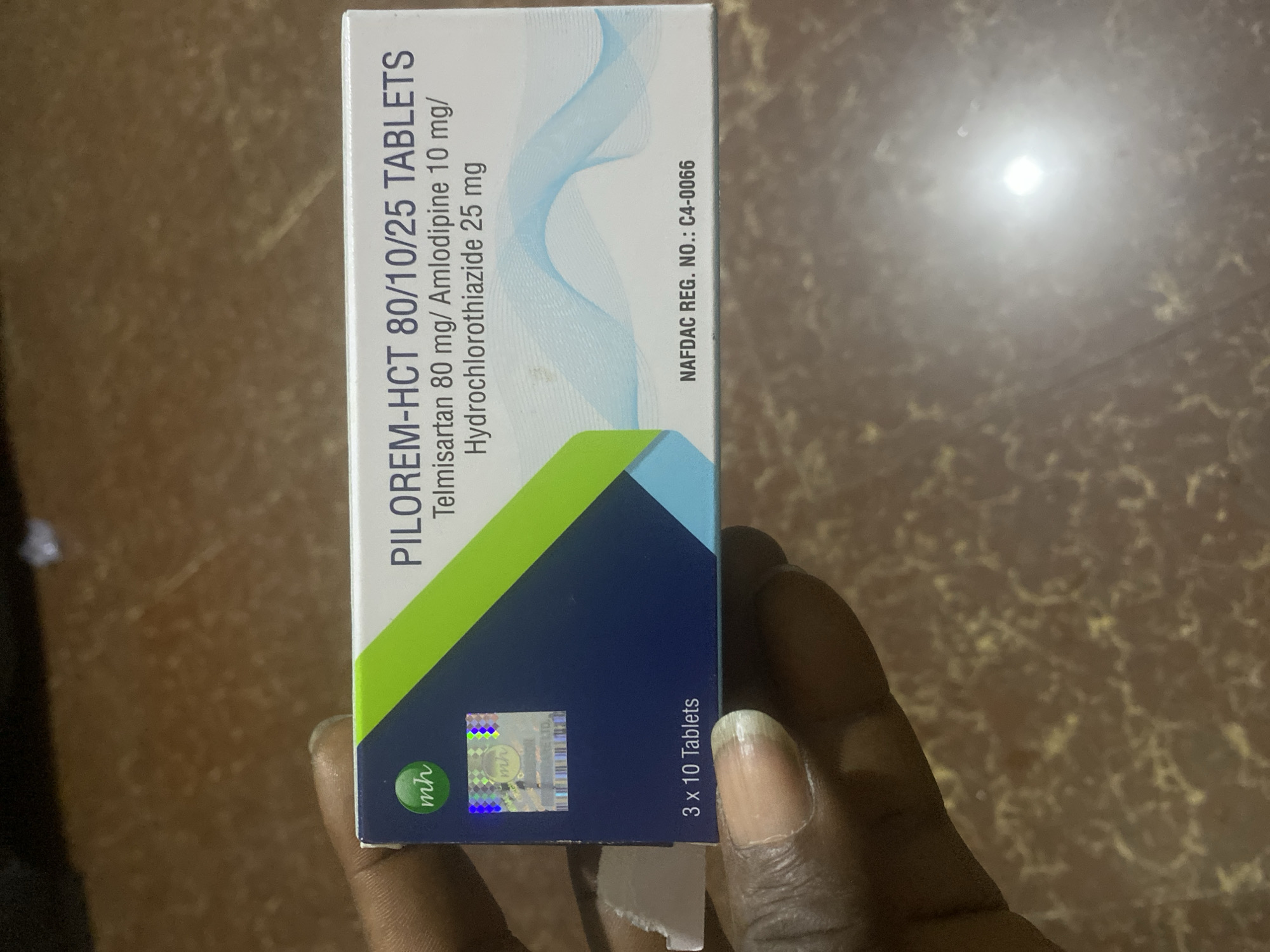 A close-up, eye-level shot reveals a dark-skinned hand holding a white and blue medicine box labeled "PILOREM-HCT 80/10/25 TABLETS" against a mottled brown background, likely a table or floor, in a location in Jos, Nigeria.
The rectangular medicine box prominently displays "PILOREM-HCT 80/10/25 TABLETS" in dark blue capital letters. Below this, the active ingredients are listed as "Telmisartan 80 mg/ Amlodipine 10 mg/ Hydrochlorothiazide 25 mg." The bottom left of the box indicates "3 x 10 Tablets," and the bottom right features "NAFDAC REG. NO.: C4-0066," signifying registration with Nigeria's National Agency for Food and Drug Administration and Control. A green "mh" logo is visible in the top left corner of the box, with a holographic sticker featuring the "mh" logo and "HEALTHCARE LTD." beneath it. The box design includes a diagonal green band across the top left, transitioning to a dark blue section, and a lighter blue, wave-like graphic on the right side.
The fingers of the hand are visible, with slightly discolored fingernails, supporting the box from underneath and to the left. The background is out of focus, showing a dark brown surface with lighter, irregular patterns, and a bright, circular light reflection in the upper right quadrant.