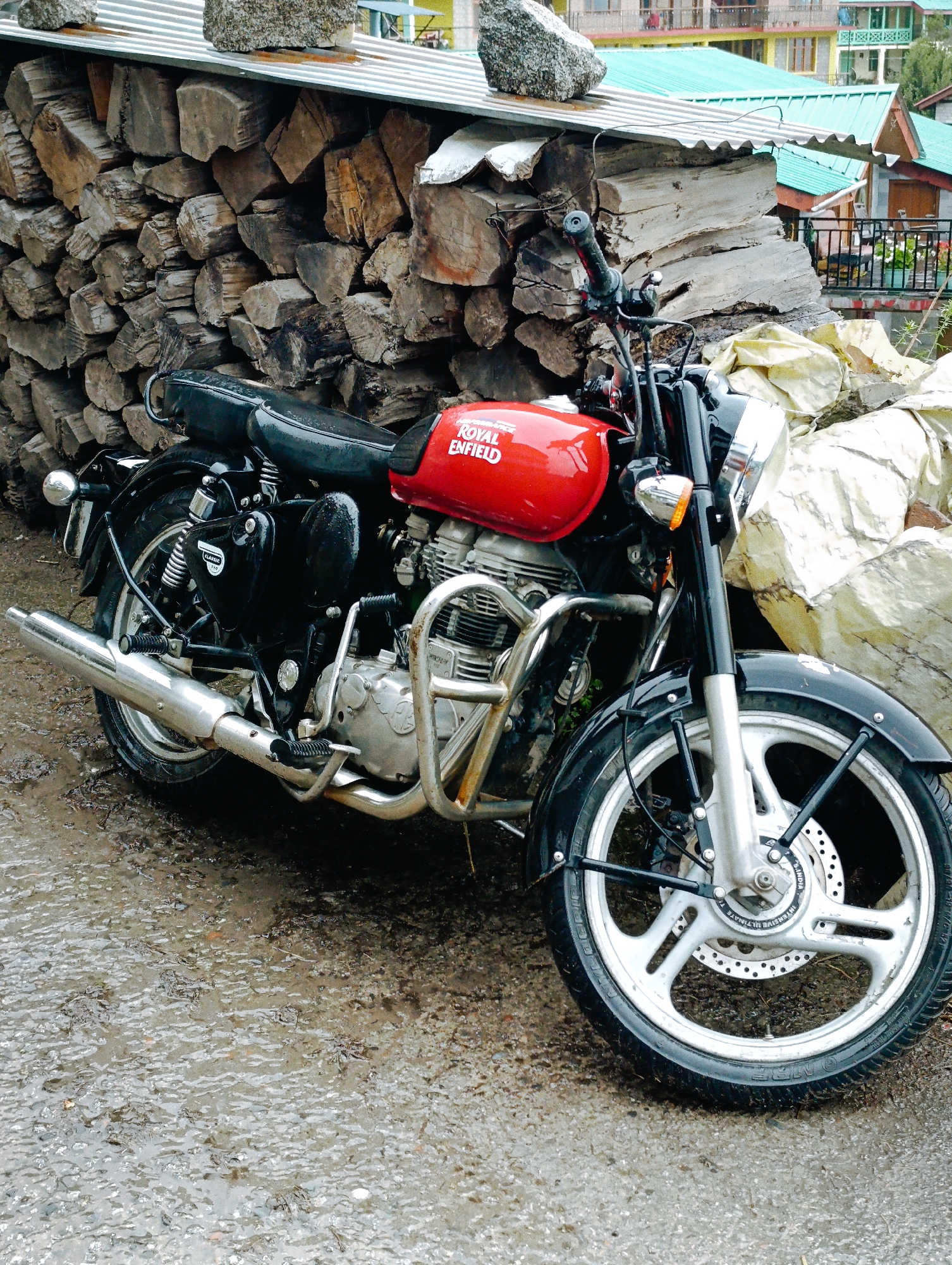 A red and black Royal Enfield motorcycle is parked on a wet, muddy surface in front of a wall of stacked firewood. The motorcycle is the primary subject, with its fuel tank prominently displaying the "ROYAL ENFIELD" logo in white lettering. The rear tire is visible on the left, and the front wheel and fender are on the right. The scene appears to be outdoors, with a building and some vegetation visible in the background. The overall impression is one of a rural or semi-urban environment in Palchan, India, possibly after a rainfall given the wet ground. No people are present in the image.