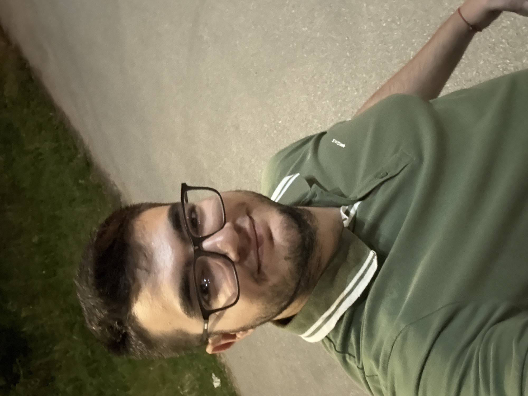 An individual is depicted in a close-up, waist-up, angled selfie taken outdoors during the evening or night. The subject is a male, appearing to be in his twenties, with dark hair, a light beard, and black-framed eyeglasses. He is smiling faintly, looking directly into the camera. He wears a dark olive-green polo shirt with a white collar featuring two thin horizontal green stripes. A small, white embroidered logo or text, "MCGAS," is visible on the upper left chest of the shirt. A thin red string bracelet is present on his left wrist. The background features a paved surface, likely a pathway or sidewalk, extending to the right and foreground. To the left, dark, out-of-focus foliage or grass is visible. The scene is illuminated by an artificial light source, casting highlights on the subject's face and glasses. The specific location within Sofia, Bulgaria, cannot be determined from the visual information provided in the image.