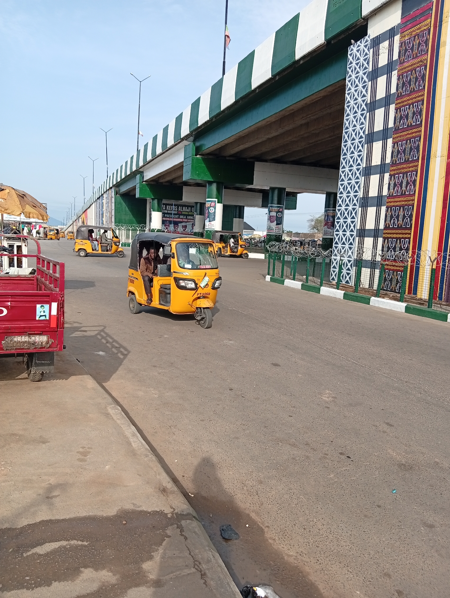The image captures a street scene in Jalingo, Nigeria, beneath an elevated highway overpass. The overpass is adorned with green and white stripes and features large, colorful decorative panels with geometric and traditional patterns. In the foreground, a yellow auto-rickshaw, also known as a Keke Napep, with the registration plate PT 906R, is driven by a man wearing a brown shirt. Another yellow Keke Napep is visible in the background. To the left, the rear of a red truck is parked. The road surface appears worn, with some debris and water stains. Fencing with barbed wire runs along the side of the overpass, and political posters are affixed to the supporting pillars. The sky is clear and blue, indicating daylight.