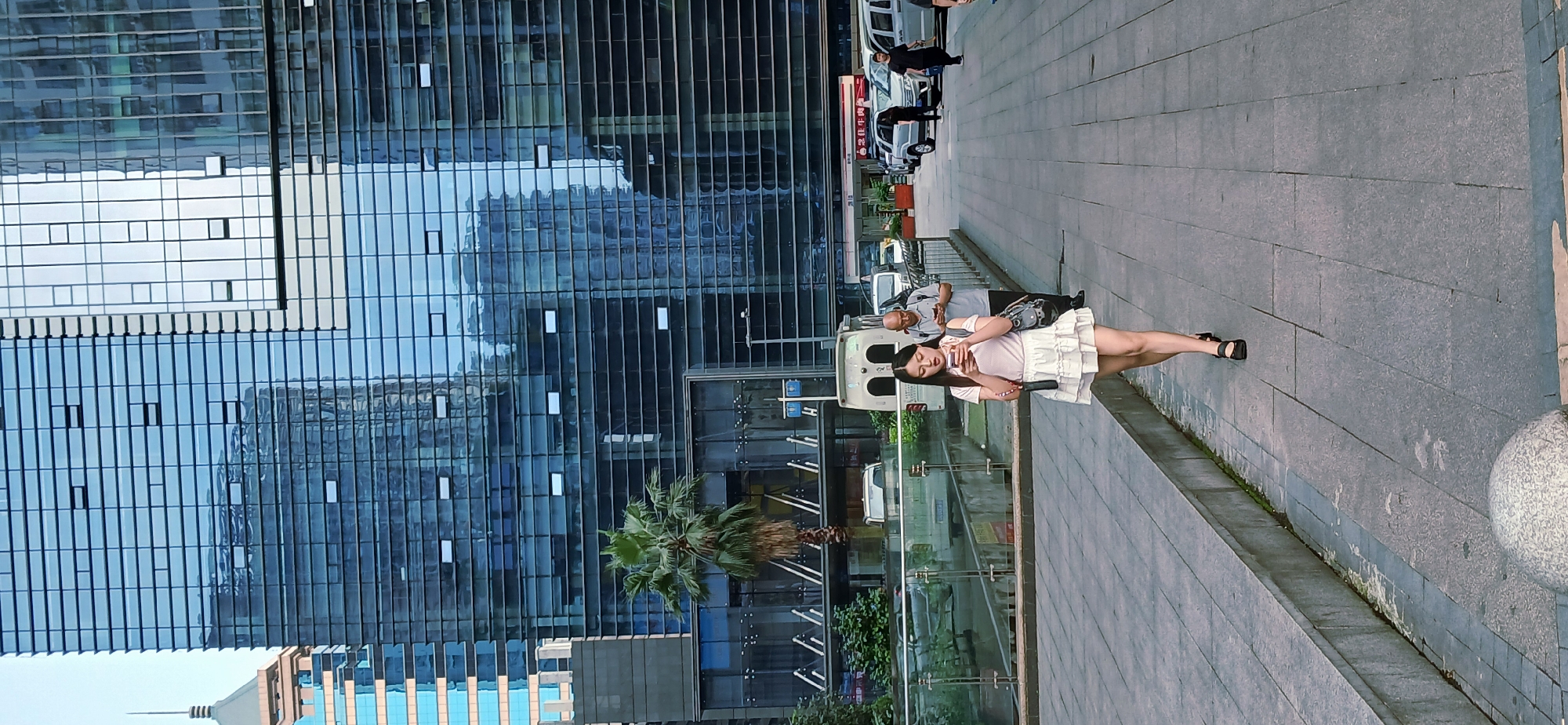 A medium, eye-level, outdoor shot depicts a female individual standing on a paved sidewalk in Jiulongpo District, China.
The woman is light-skinned with long, dark hair. She is wearing a light-colored top and a short, white, tiered skirt. She holds a dark-colored object, possibly a phone, in her hand and appears to be looking at it. She wears black sandals.
Behind the woman, a modern building with a glass facade dominates the background. The glass reflects the sky and the surrounding environment. To the side of the building, a palm tree is visible.
The sidewalk is made of gray tiles. In the background, vehicles are parked along the street, and several pedestrians are visible. A glass barrier separates the sidewalk from the street. A stone barrier is to the right of the sidewalk.