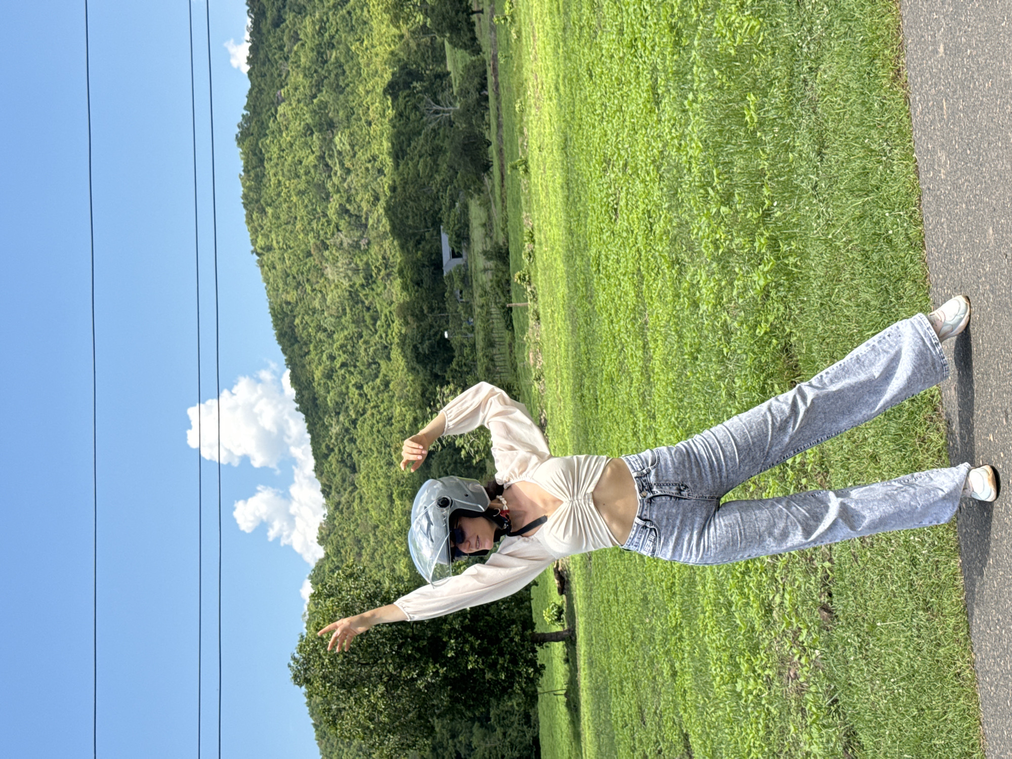 A woman wearing a light-colored, ruched crop top, acid-wash wide-leg jeans, and white sneakers stands on an asphalt path. She has on a silver motorcycle helmet with the visor up, sunglasses, and a red and white bandana tied around her neck. Her left arm is raised with her hand open, palm facing outward, and her right arm is bent at the elbow, with her hand held near her chest. She is positioned in front of a vibrant green, grassy field. In the background, a dense, lush green forest covers a hillside under a bright blue sky with scattered white clouds. Power lines are visible across the top of the frame, and a solitary tree stands to the left of the woman. The scene appears to be in a rural or semi-rural area, potentially near the city of Ham Thuan Bac, Vietnam, given the tropical vegetation and terrain.