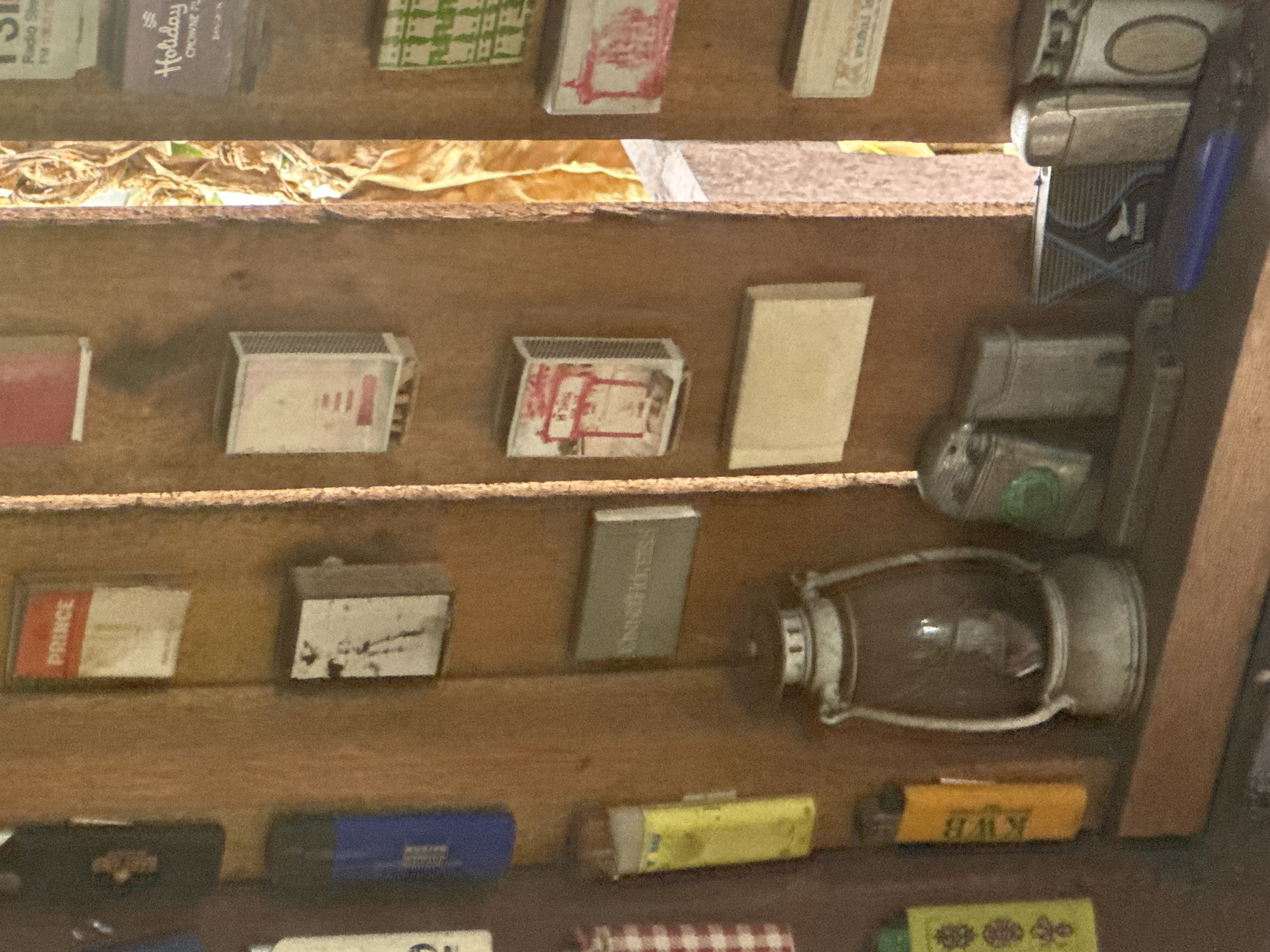 The image depicts a shelf in Baan Hua Thanon, Thailand, containing a collection of various small objects. These include multiple matchboxes with assorted designs, several lighters, and a small antique lantern with a metallic finish. The shelf itself appears to be made of wood. The lighting suggests an indoor environment, possibly within a shop or storage space.