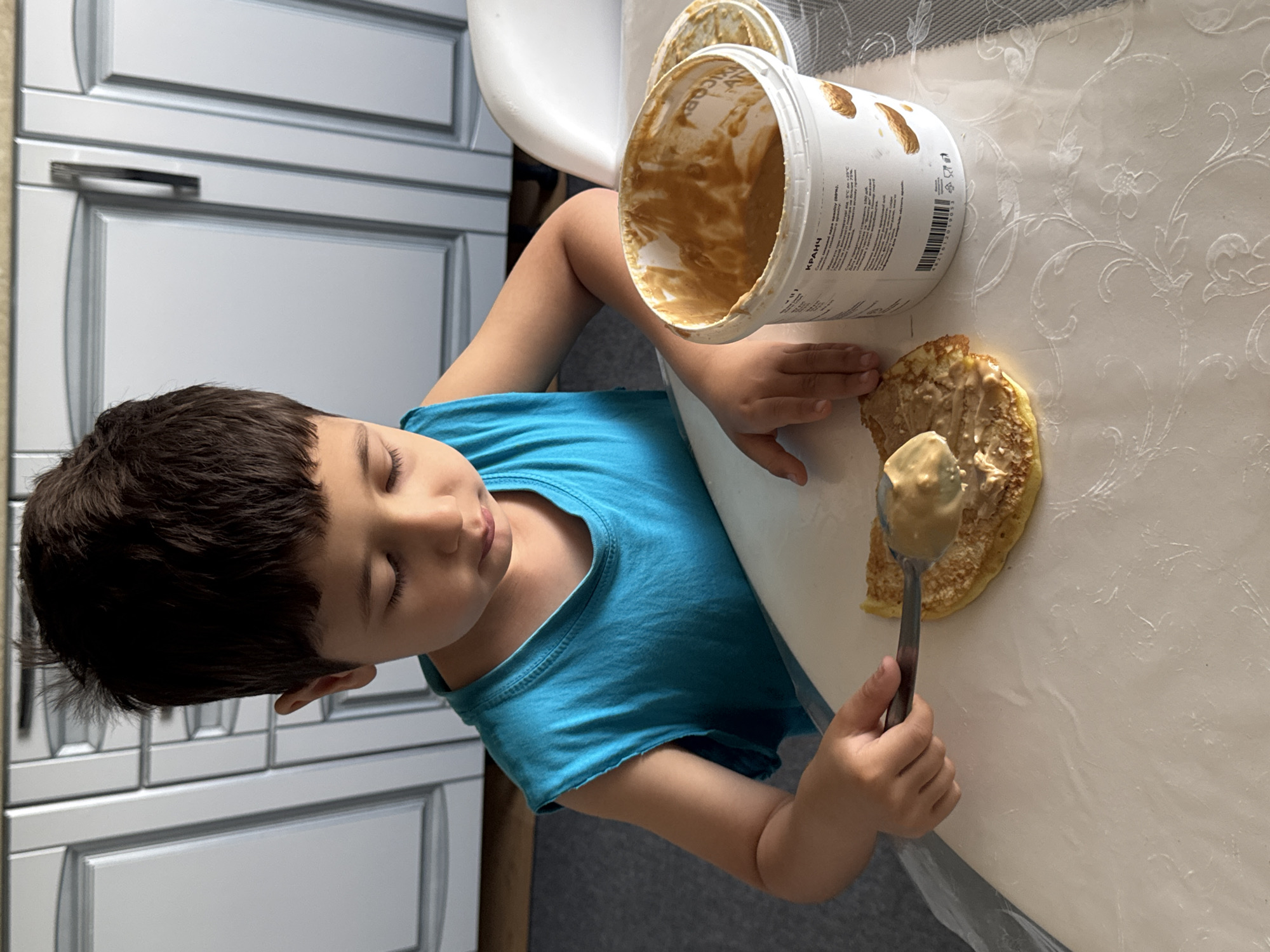 The image shows a young boy with dark hair and closed eyes, wearing a blue t-shirt. He is in a kitchen, with a white table in the foreground covered by a tablecloth with a floral pattern. To the boy's right, there is a large white tub of peanut butter, with some of the spread visible on the inside rim. A spoon is in the boy's right hand, holding a dollop of peanut butter, and he appears to be in the process of spreading it onto a pancake that is lying on the table. The boy's expression is serene, suggesting concentration or enjoyment in his task. In the background, to the left of the frame, are white kitchen cabinets with silver handles. The lighting suggests it is daytime. There is text on the peanut butter tub, but it is not clearly legible.